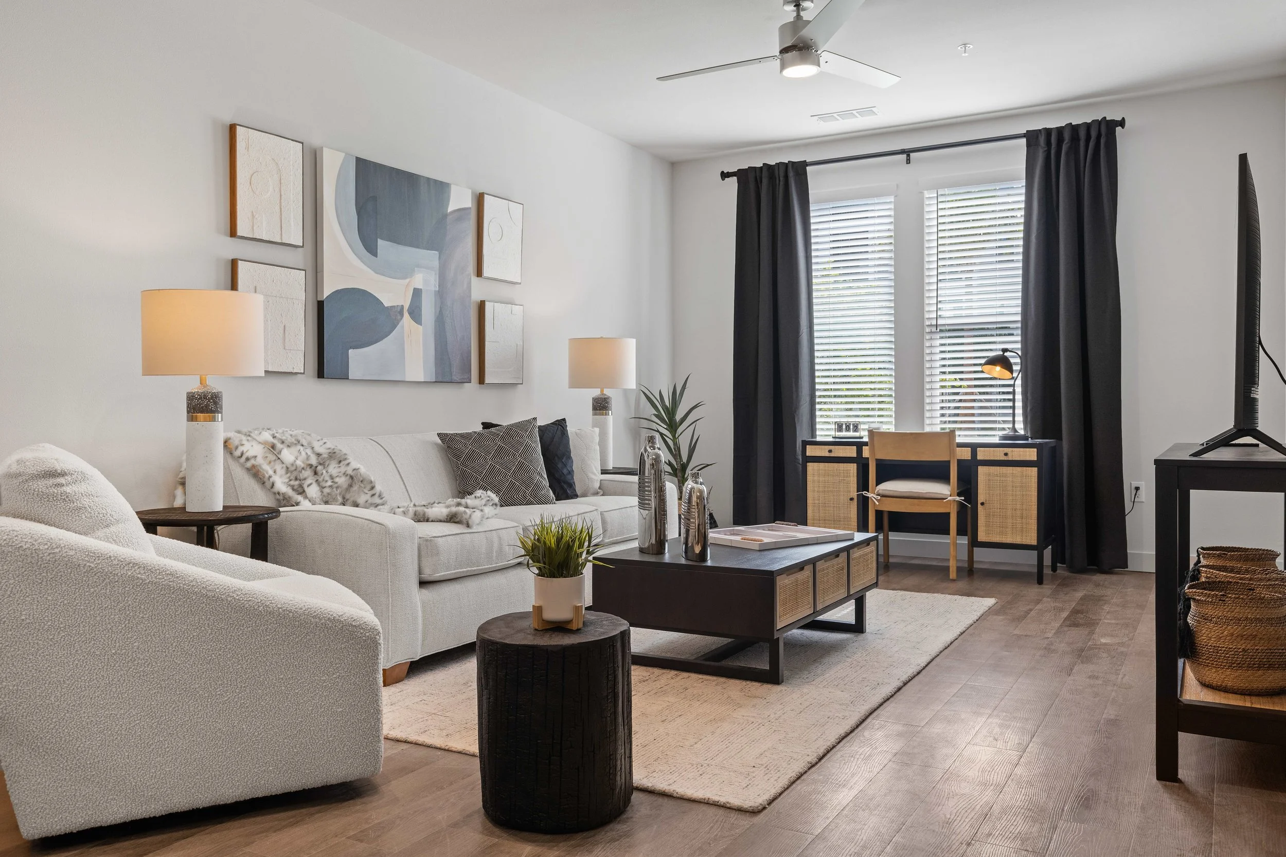 A modern living room with white couches, dark wood coffee table, abstract artwork on the wall, beige and black accents, a side table with lamps, large windows with white blinds and black curtains, ceiling fan, TV, and decorative plants.