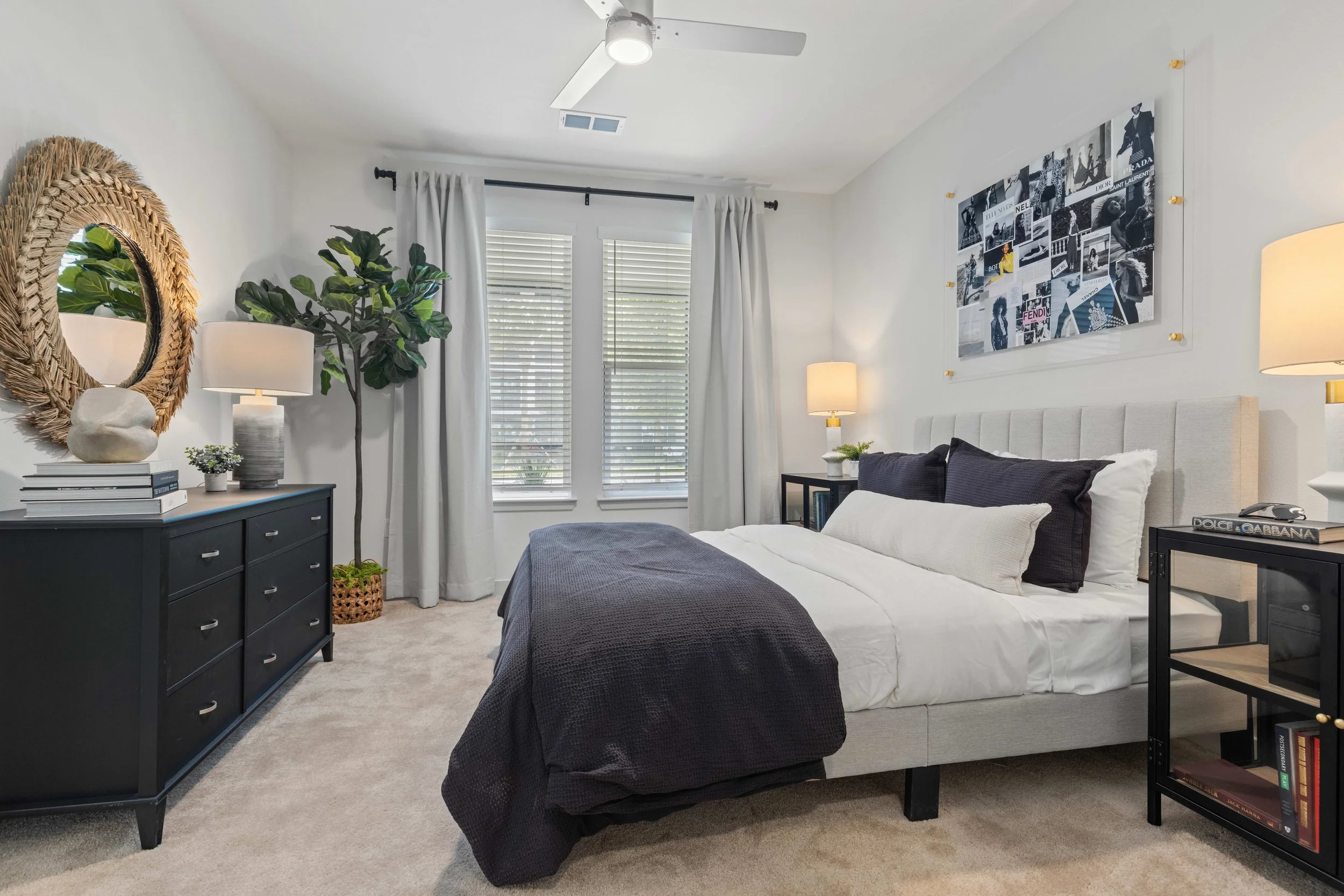 A bedroom with white walls, two large windows with white blinds and curtains, a beige upholstered headboard, black and white artwork on the wall, a black dresser with decorative items, two cream-colored lamps, a green plant, and a mix of black, cream