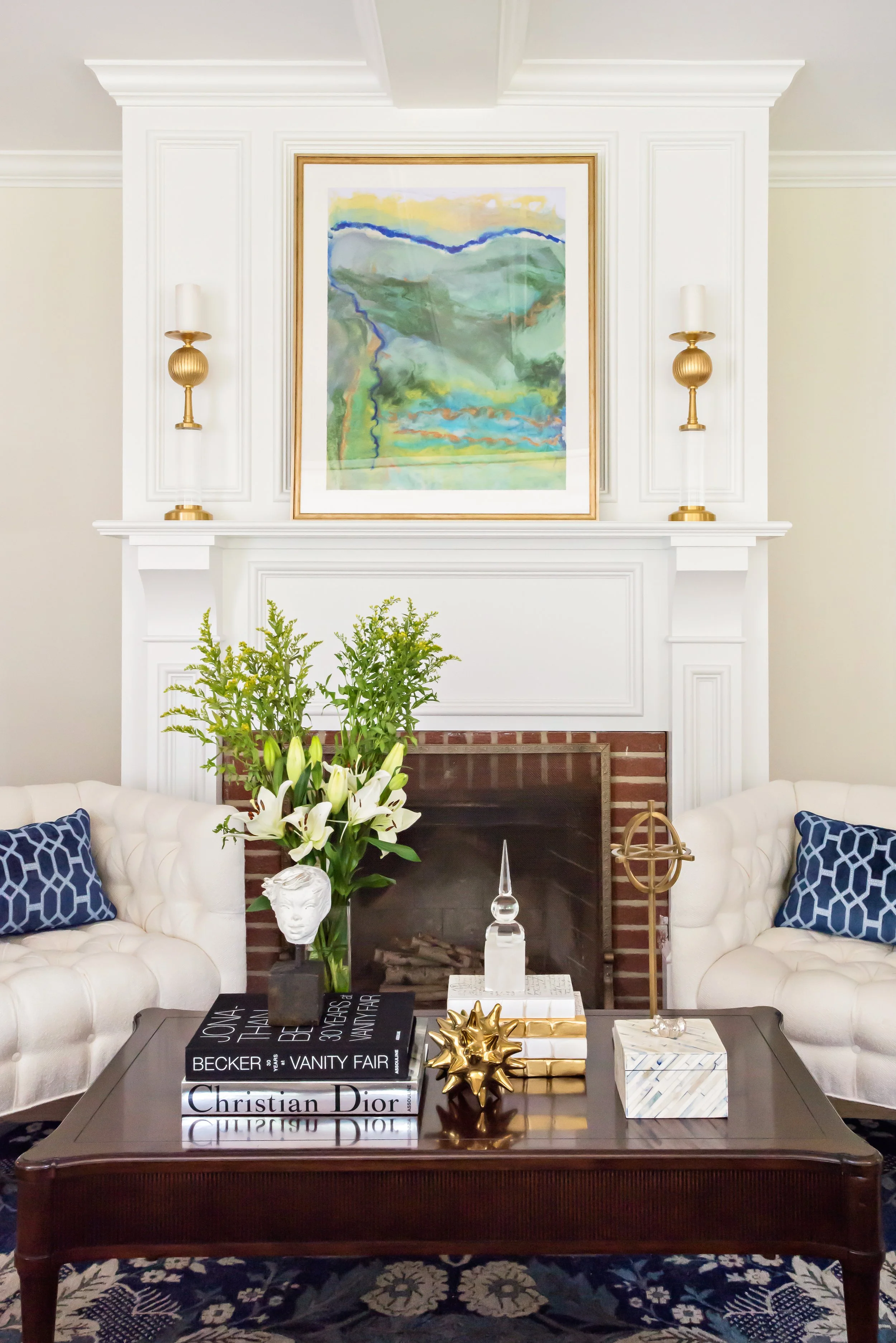 Living room with a white fireplace, landscape painting above, two gold sconces, white sofas with blue patterned pillows, centerpiece with white lilies and greenery, coffee table with fashion books, gold decorative objects, and a marble box, rug with 