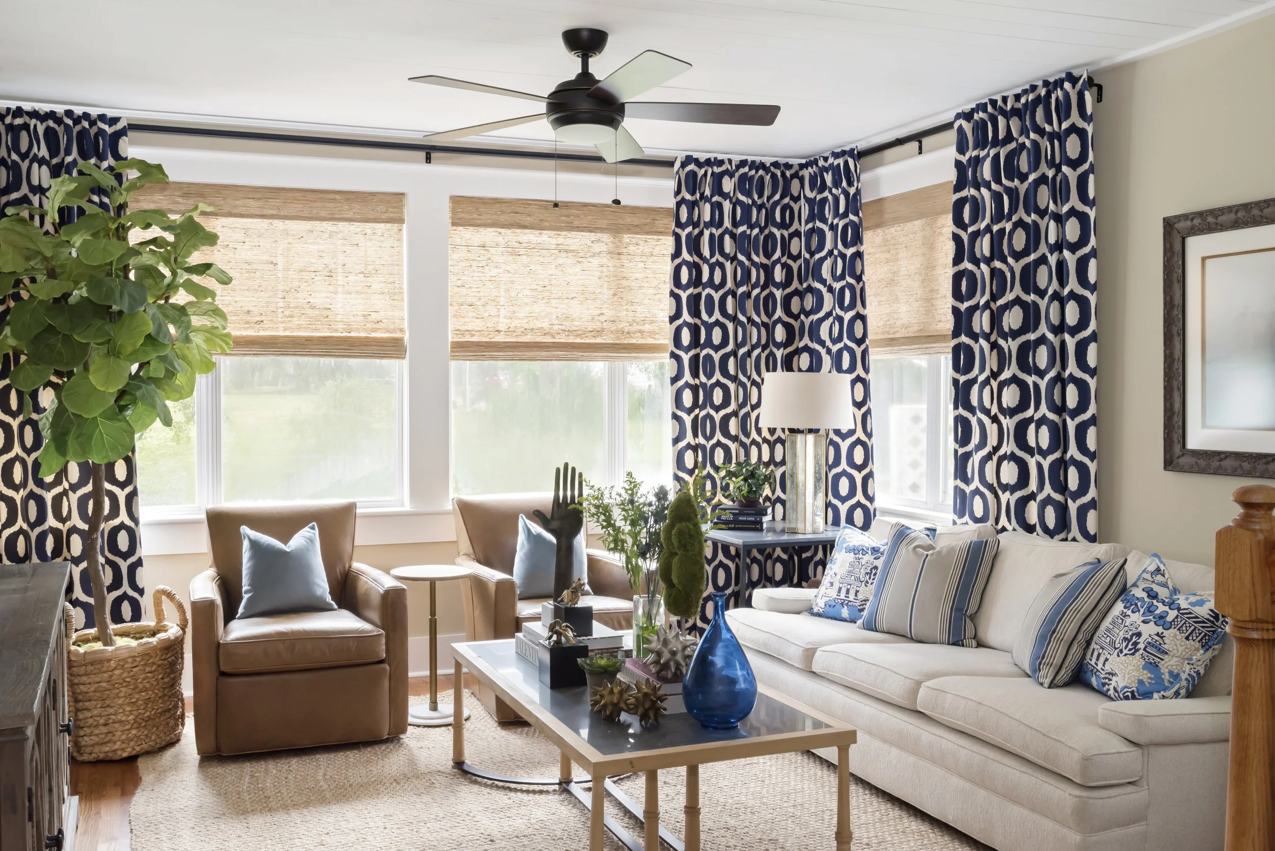 A living room with a ceiling fan, large windows with bamboo blinds and blue patterned curtains, a beige sofa with decorative pillows, a wooden coffee table with blue vases and decorative objects, a table lamp, and several potted plants.