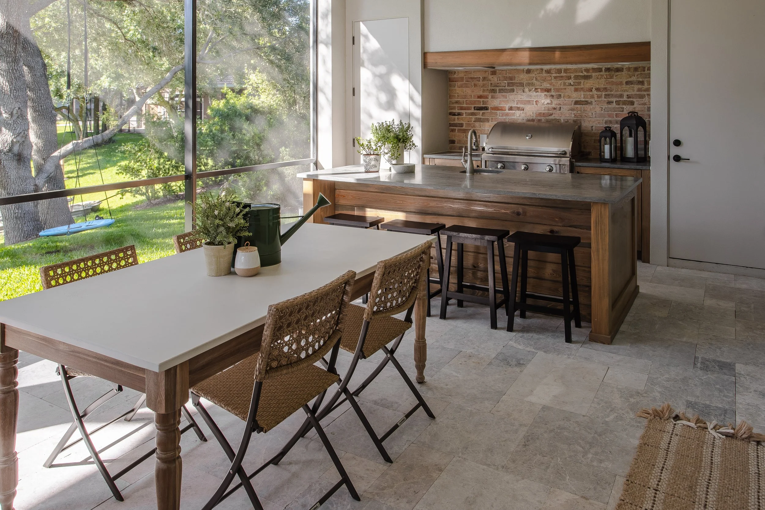 A cozy kitchen with an open layout, featuring a white dining table with woven chairs, a stone and wood countertop island with bar stools, a stainless steel grill, and a large window opening to a green outdoor garden with trees and a swing.