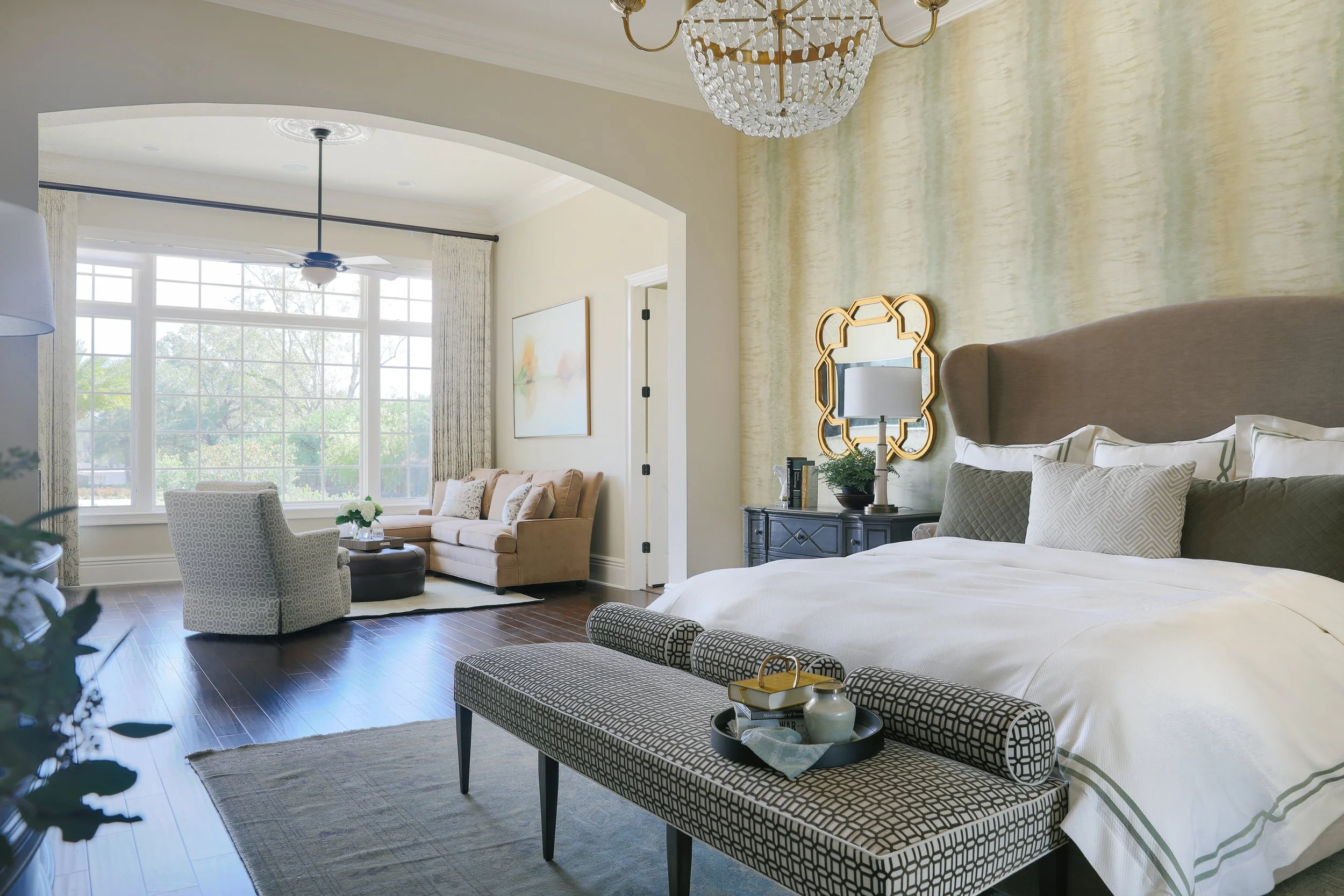 A spacious bedroom with a large bed, decorative pillows, and a patterned bench at the foot of the bed. There is a black dresser with a lamp and mirror above it. The wall behind the bed has a textured yellow wall treatment. To the left, a sitting area