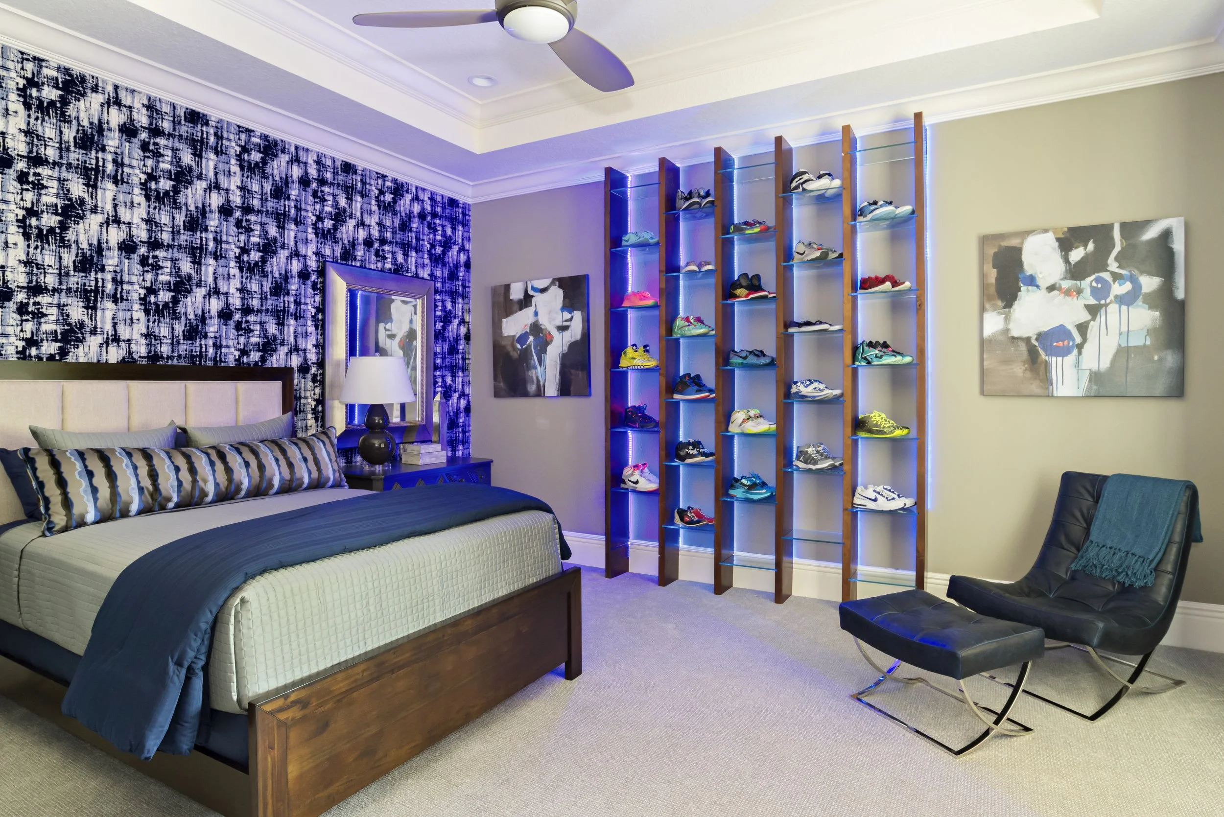 A bedroom with a bed, nightstand, and table lamp on the left. A wall with abstract black and white artwork adjacent to a shelving unit filled with sneakers, illuminated with blue LED lights. A ceiling fan is visible above. On the right, there is a bl
