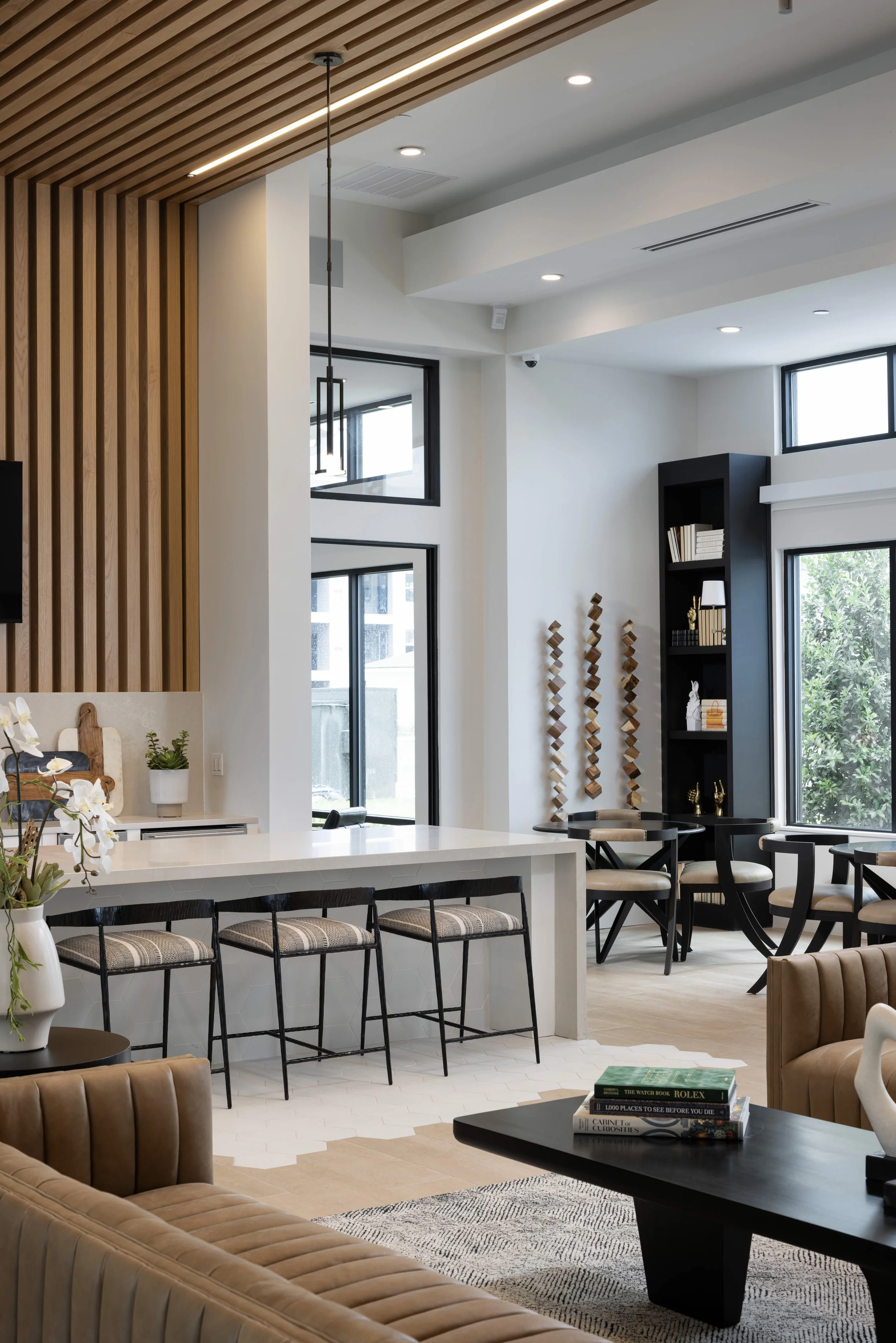 Modern indoor living space with a kitchen island, bar stools, a black dining table with chairs, a black bookshelf with decorative items, large windows, and wooden ceiling accents.