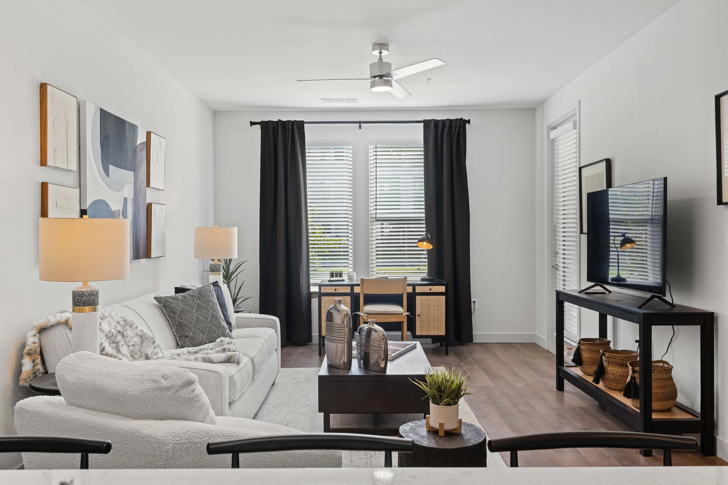 A modern living room with a white sofa, black and white patterned pillows, glass vases on a black coffee table, and a TV on a black stand. The room has white walls with abstract art and a large window with black curtains.