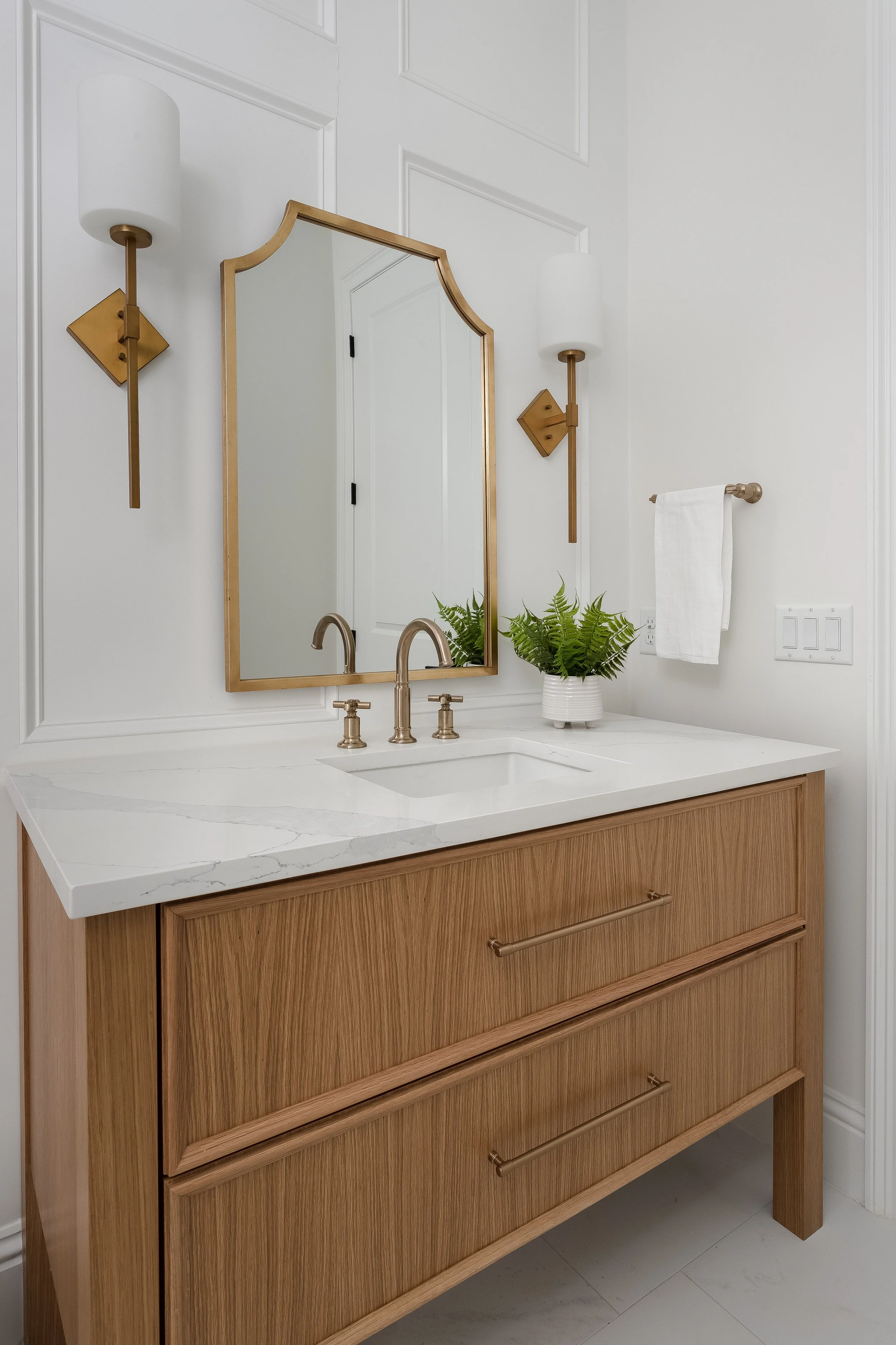 A bathroom vanity with a wooden cabinet, white marble countertop, brass faucet, mirror, wall sconces, a plant in a white pot, and a towel rack with a white towel.
