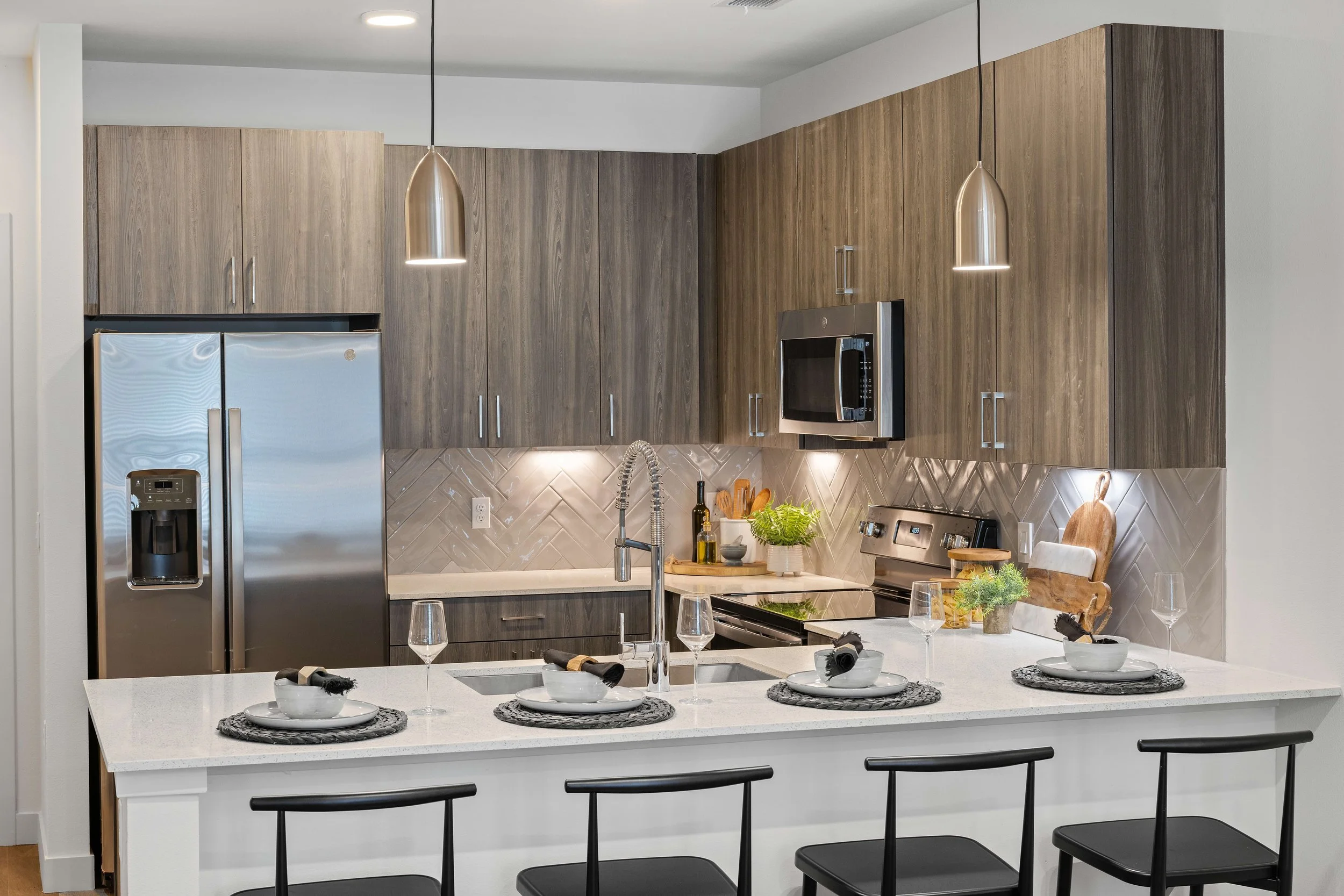Modern kitchen with a white island, black chairs, wooden cabinets, stainless steel refrigerator and microwave, pendant lights, and a kitchen countertop with plates, bowls, glasses, and greenery.
