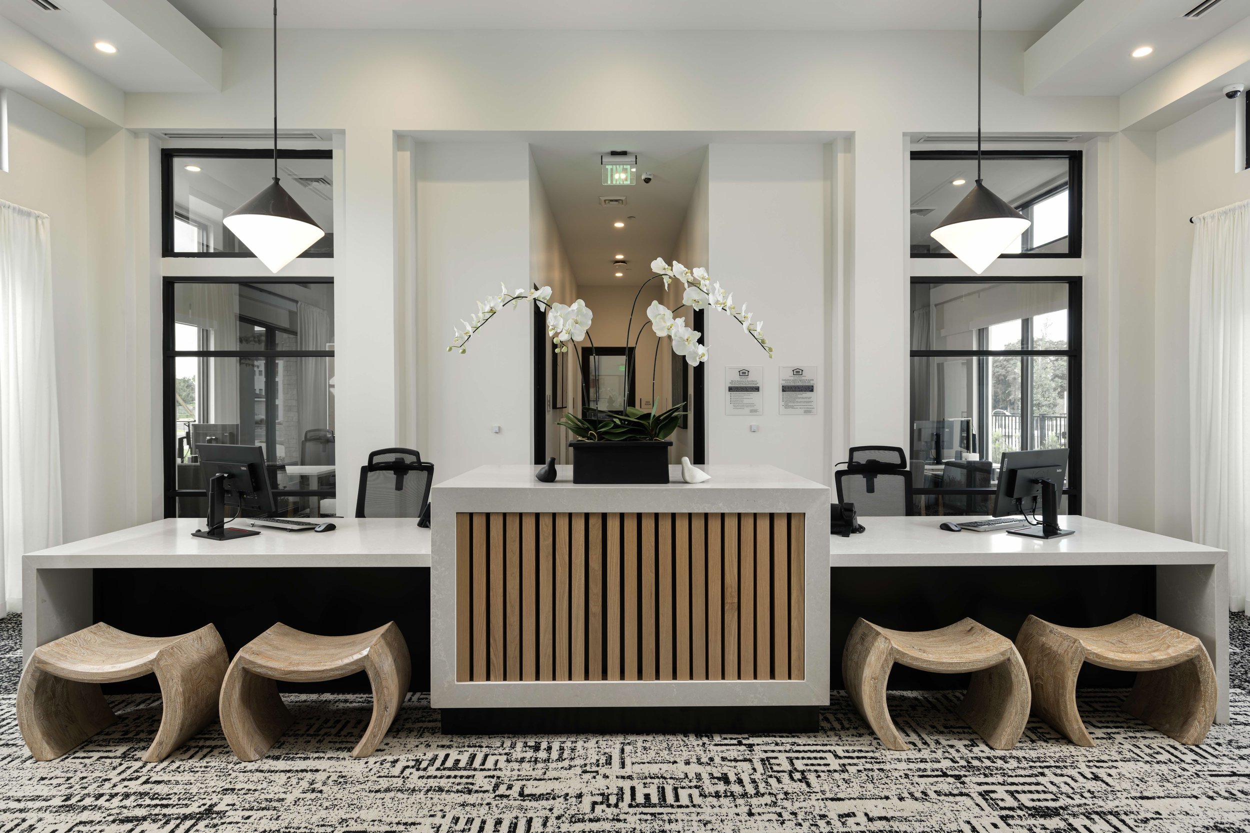 Modern office reception area with a white counter, orchid plant centerpiece, and wooden stools, flanked by computer workstations and large windows.