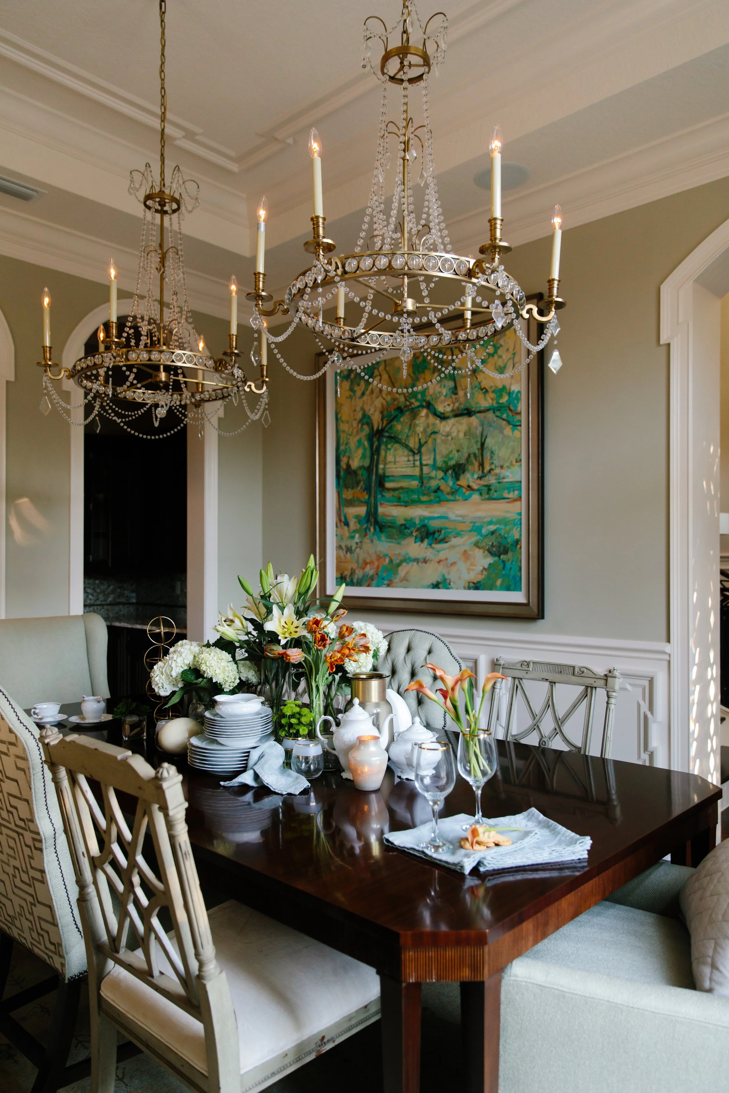 A dining table set for a meal with floral arrangements, white dishes, silverware, glasses, and a white cloth napkin. The table is surrounded by upholstered and wooden chairs. Above the table, two ornate chandeliers with lit candles and hanging crysta