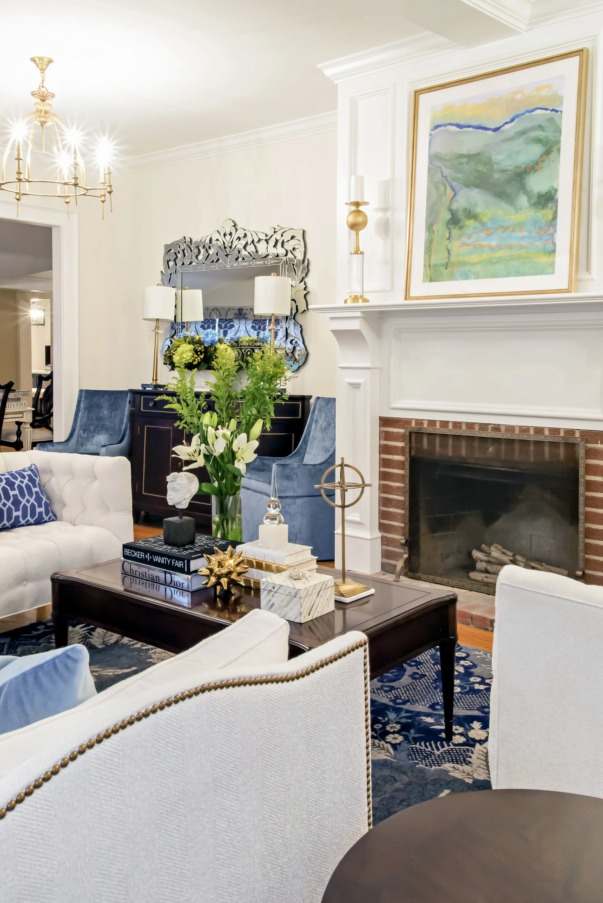 Living room with white sofa, dark wood coffee table, blue chairs, floral arrangements, fireplace, and artwork.