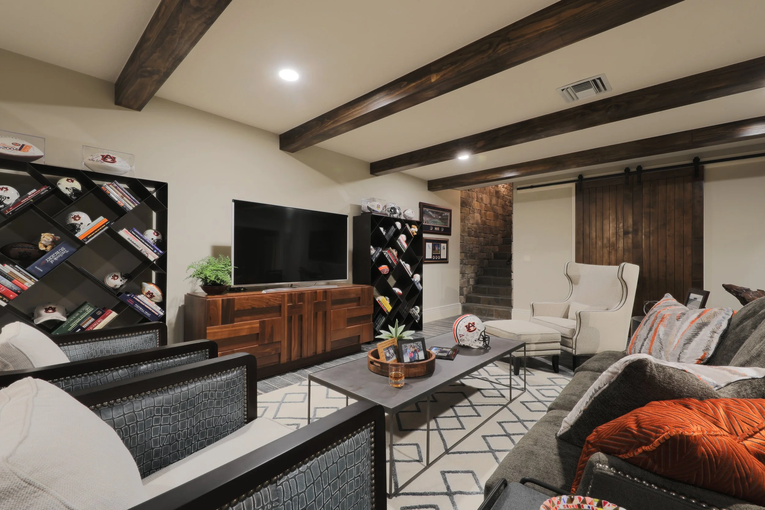 Living room with beige walls, dark wood ceiling beams, a large flat-screen TV on a wooden cabinet, black bookshelves with football helmets and books, a glass-top coffee table with framed photos, patterned rug, beige armchair, and a dark sofa with pil