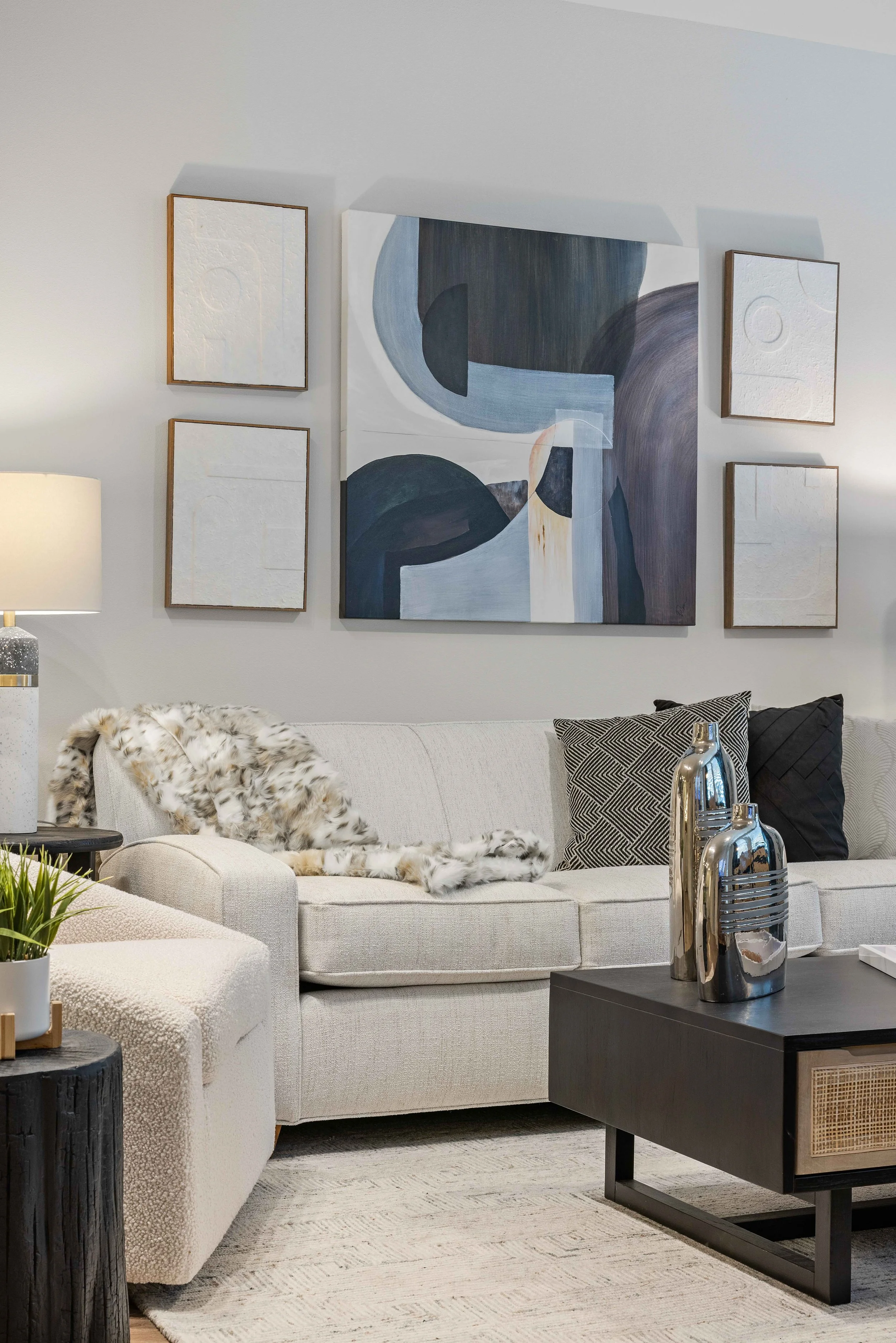 Living room with white sofa, black coffee table with metal vases, wall art with abstract shapes, and a lamp, with fur throw and patterned pillows.
