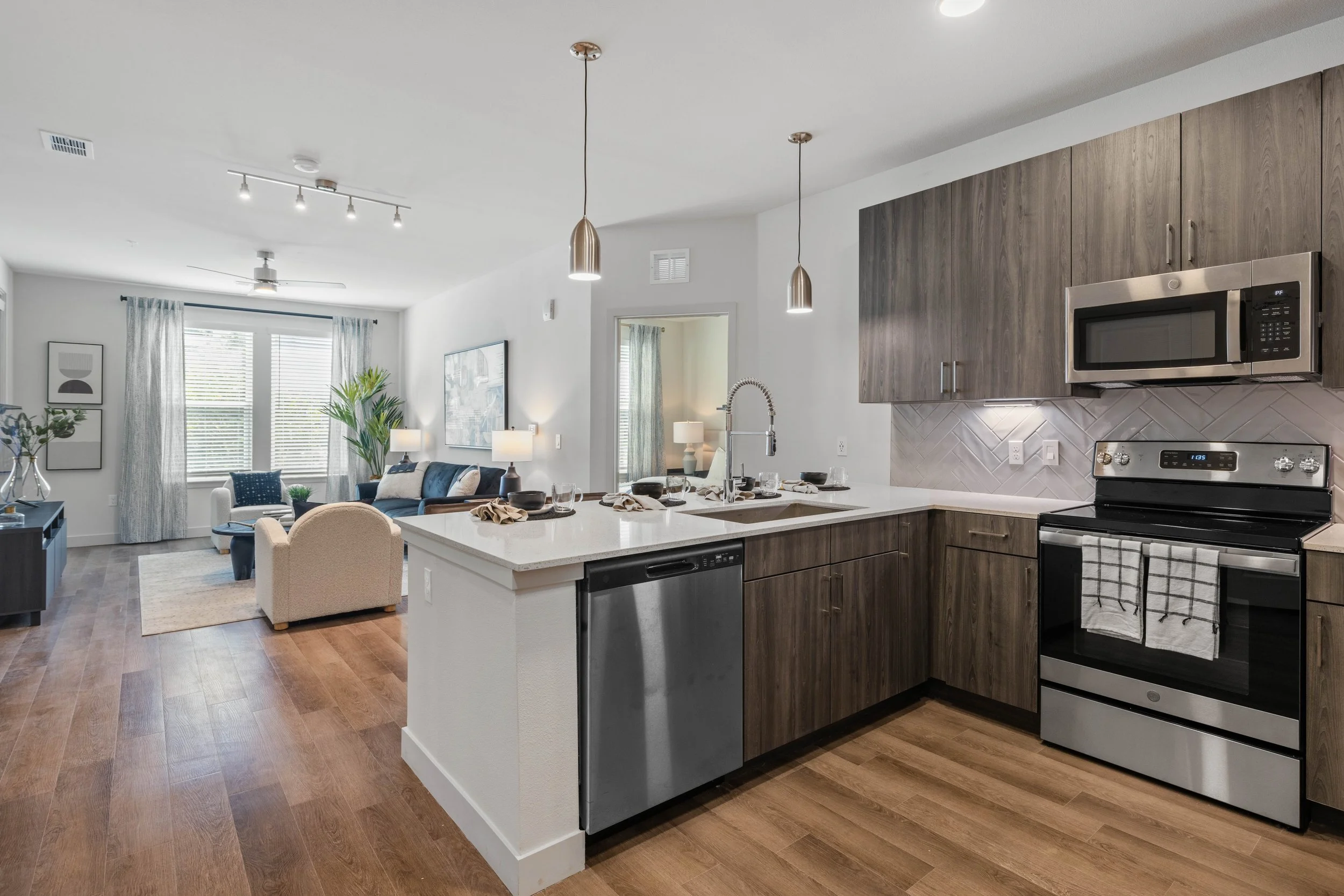 Open-concept living room and kitchen with wooden flooring, white walls, hanging pendant lights, a ceiling fan, a sofa with accent pillows, a coffee table, a dining table with chairs, large windows with curtains, and kitchen cabinets with stainless st