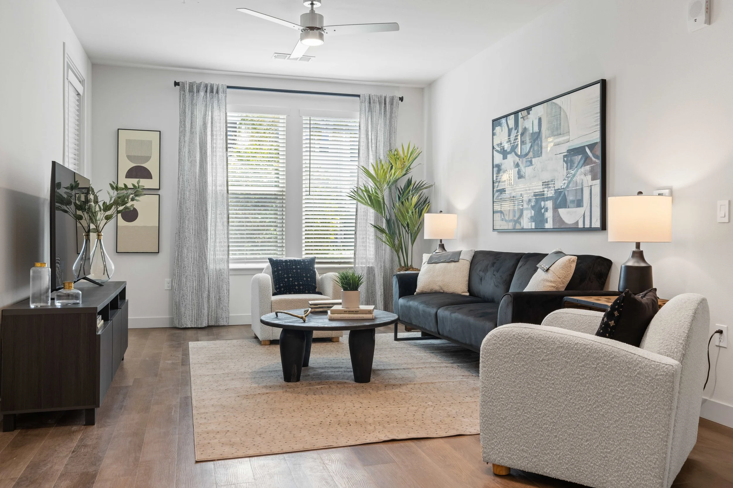 Living room with a gray sofa, beige armchair, black coffee table, wooden flooring, white walls, large window with blinds and curtains, ceiling fan, plants, wall art, and lamps.