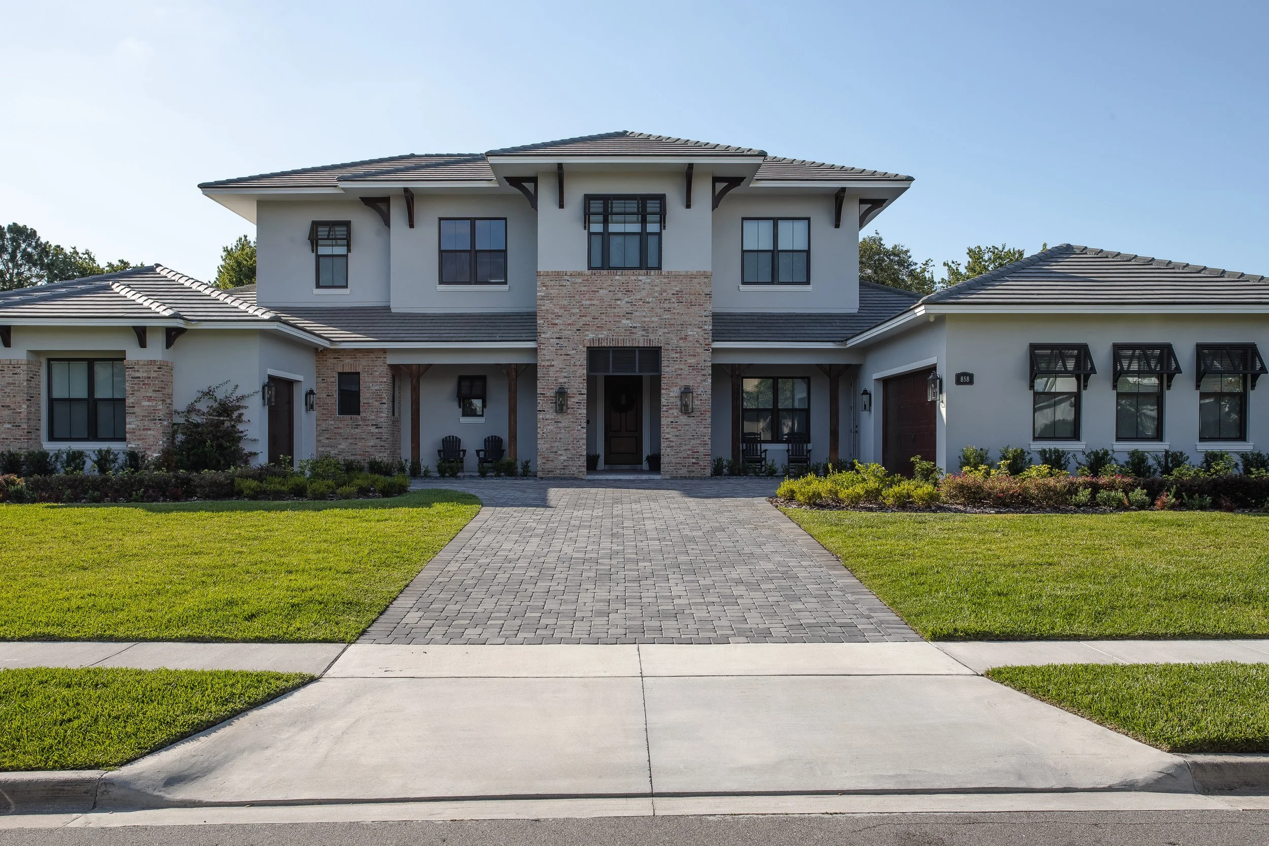 Front view of a modern two-story house with a brick and white exterior, black window frames, and a hipped roof, with a paved walkway leading to the front door, surrounded by a green lawn and landscaping.