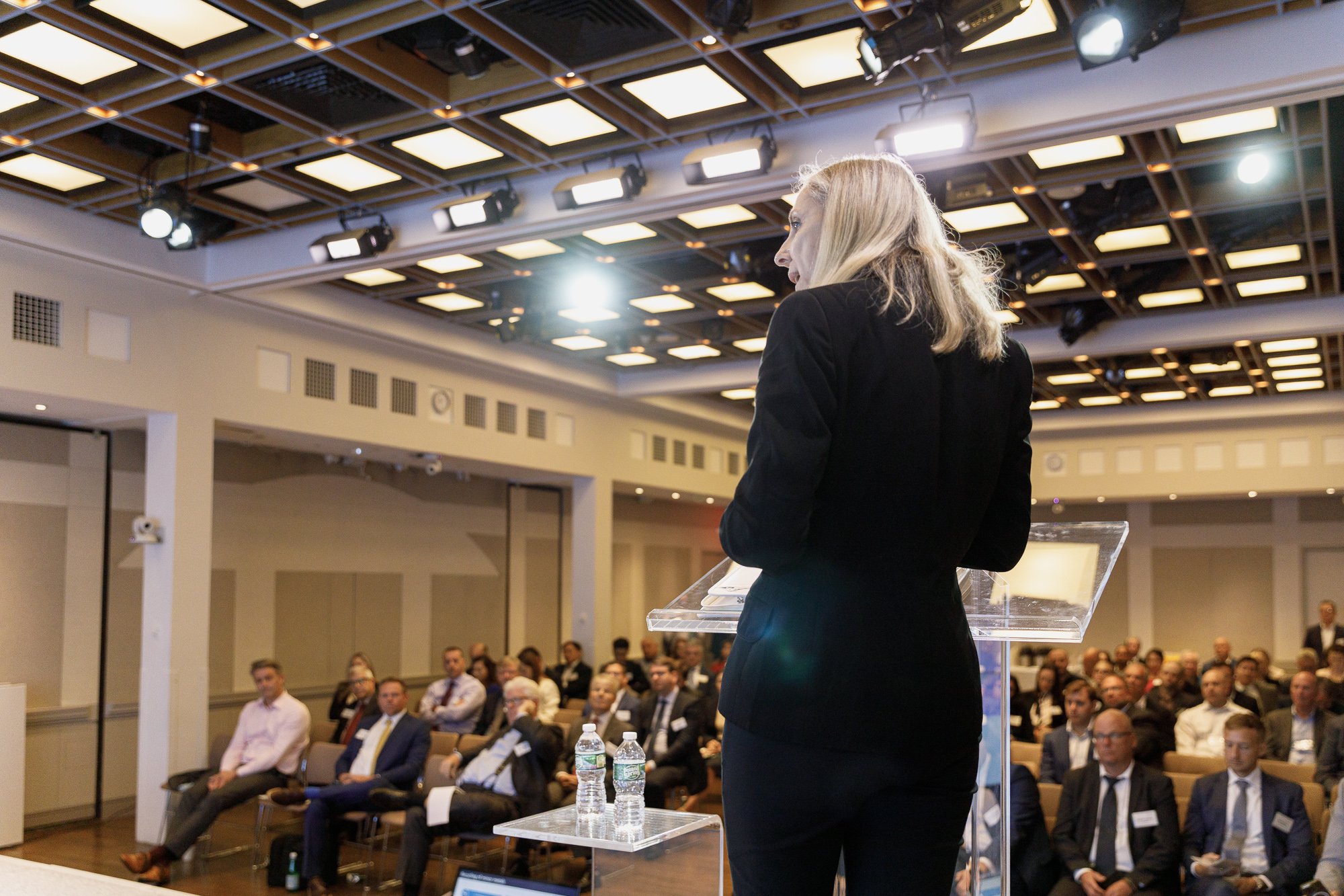 A woman in a black suit speaking at a podium during a conference with an audience seated in a large, well-lit room.
