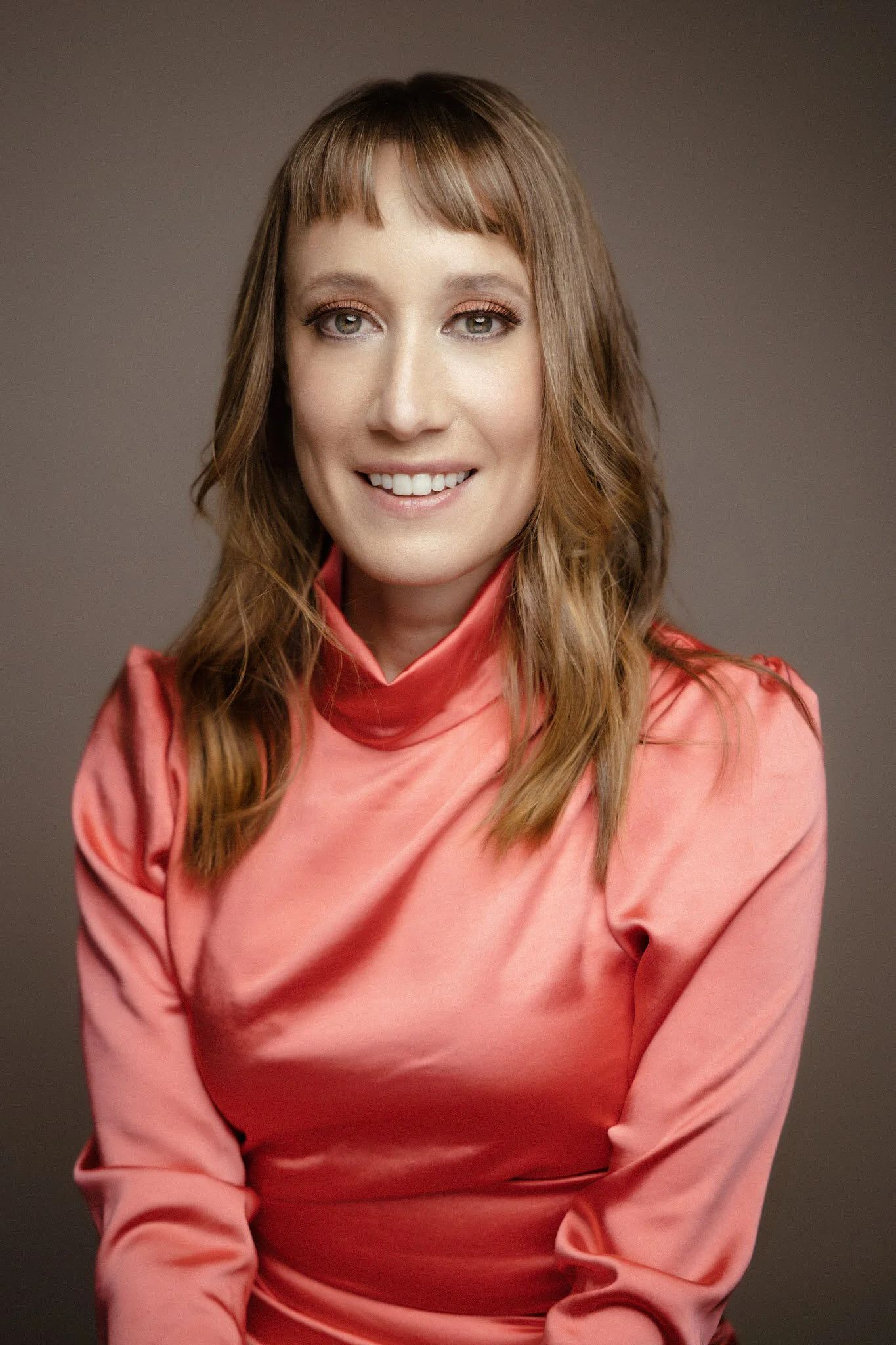 A woman with shoulder-length reddish-brown hair, wearing a pink satin blouse, smiling, with a neutral gray background.