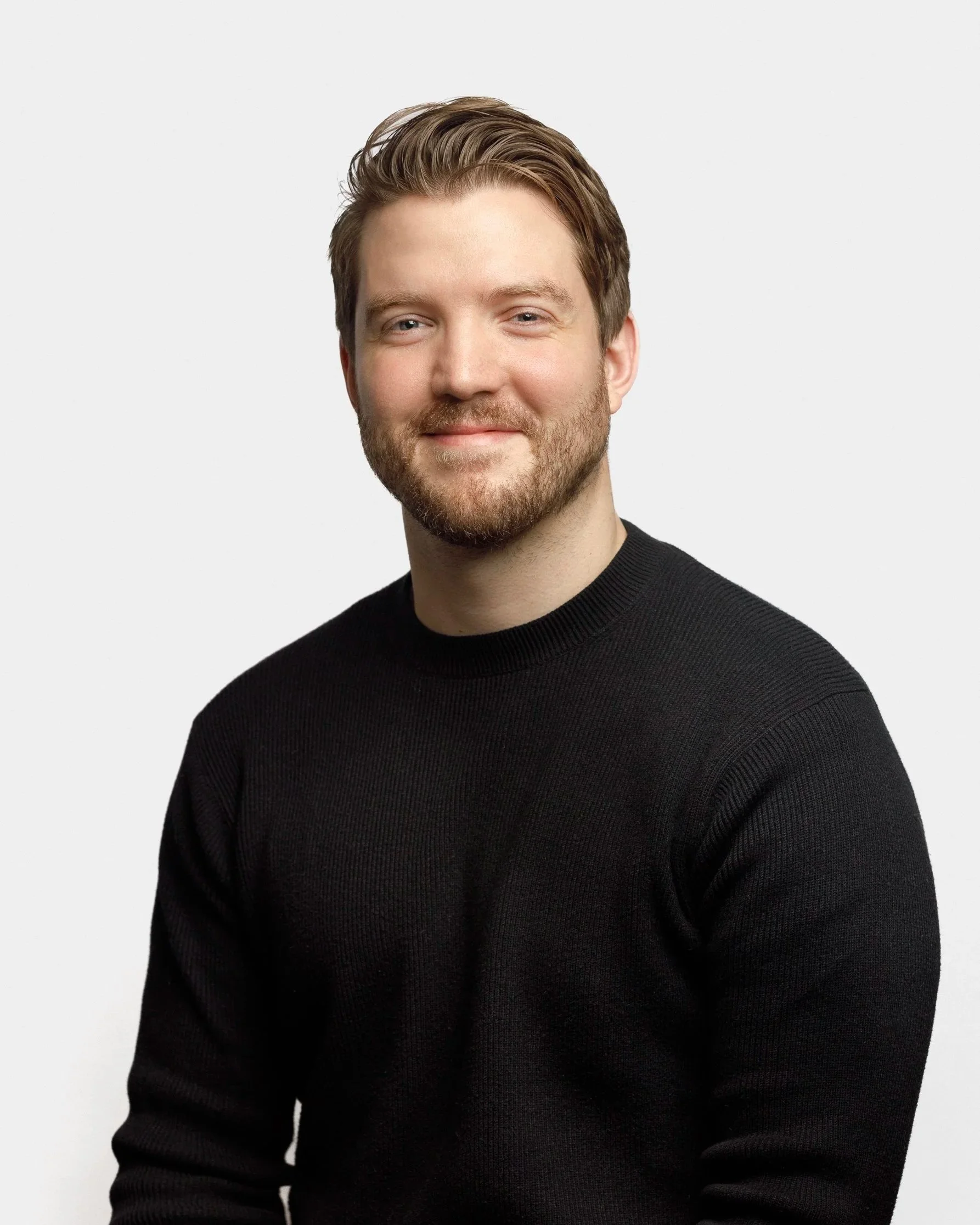 Portrait of a young man with light brown hair and a beard, wearing a black sweater, smiling against a plain light gray background.