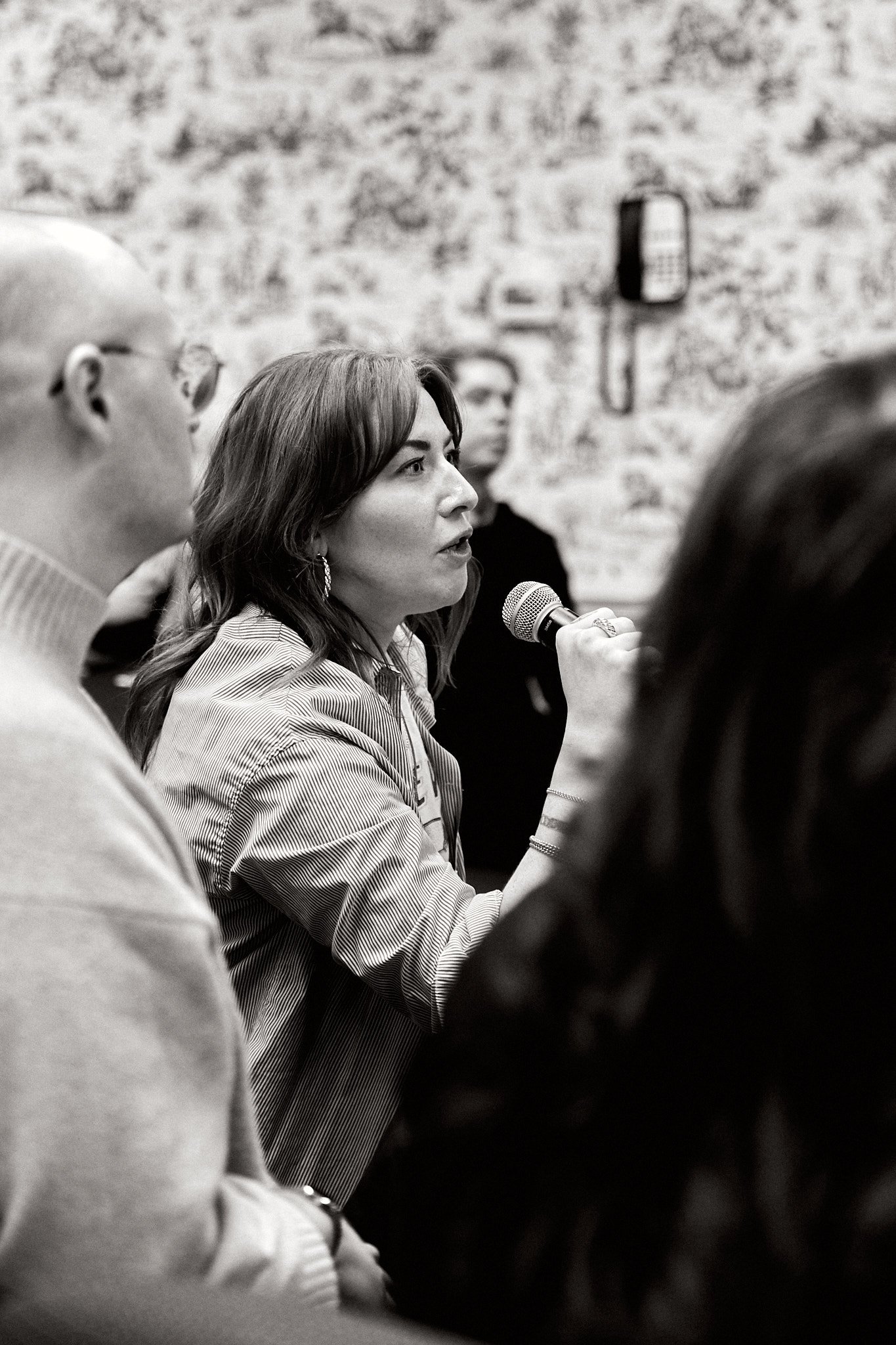 A woman is speaking into a microphone during a discussion or interview, with other people listening, in a room with a textured wall background.