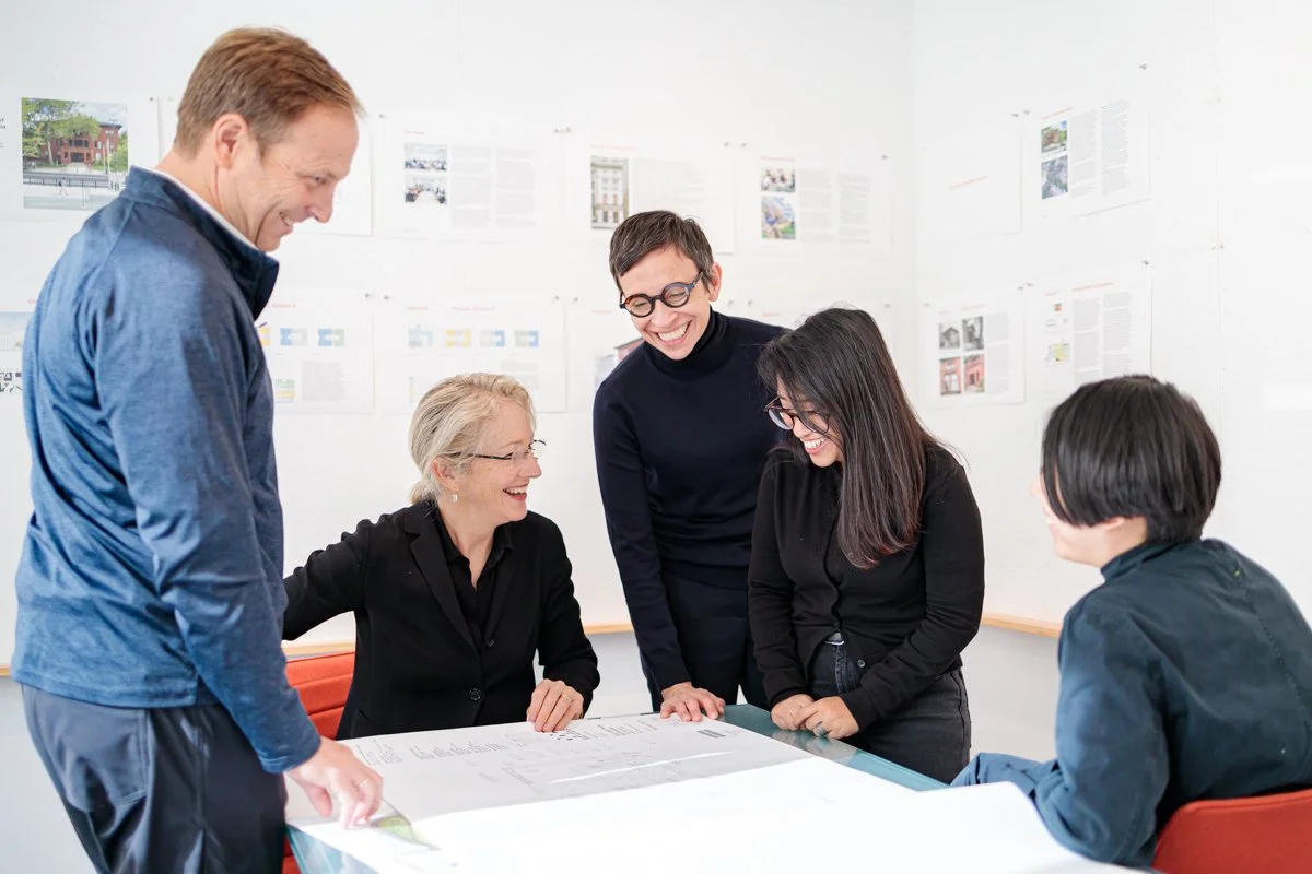 MBB Architects Team Photo in their office, Five diverse professionals, three women and two men, are gathered around a table in a bright office, sharing a laugh and discussing documents.
