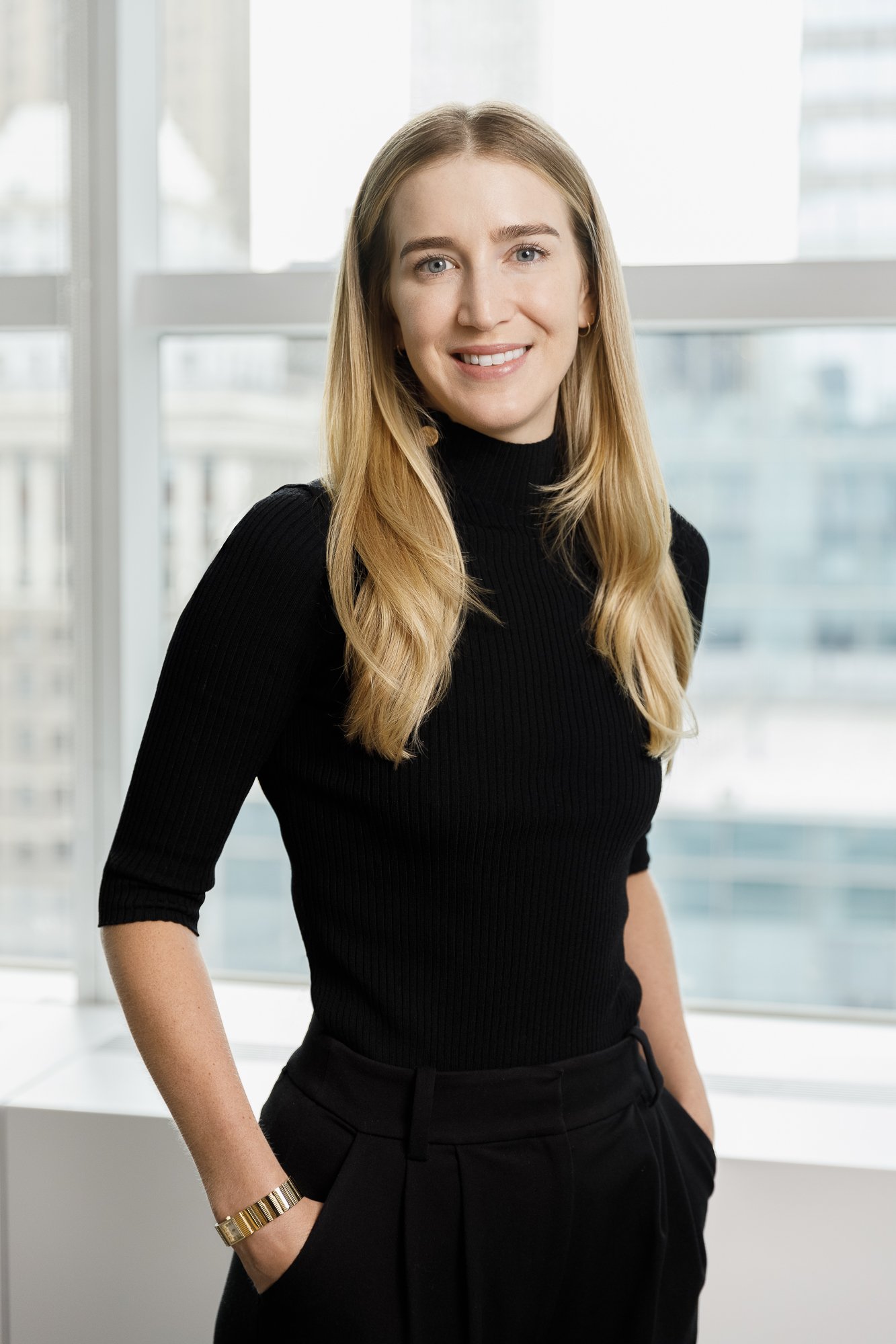 Portrait of a young woman with blonde hair wearing a black turtleneck and black pants, smiling in front of a window with a cityscape in the background.