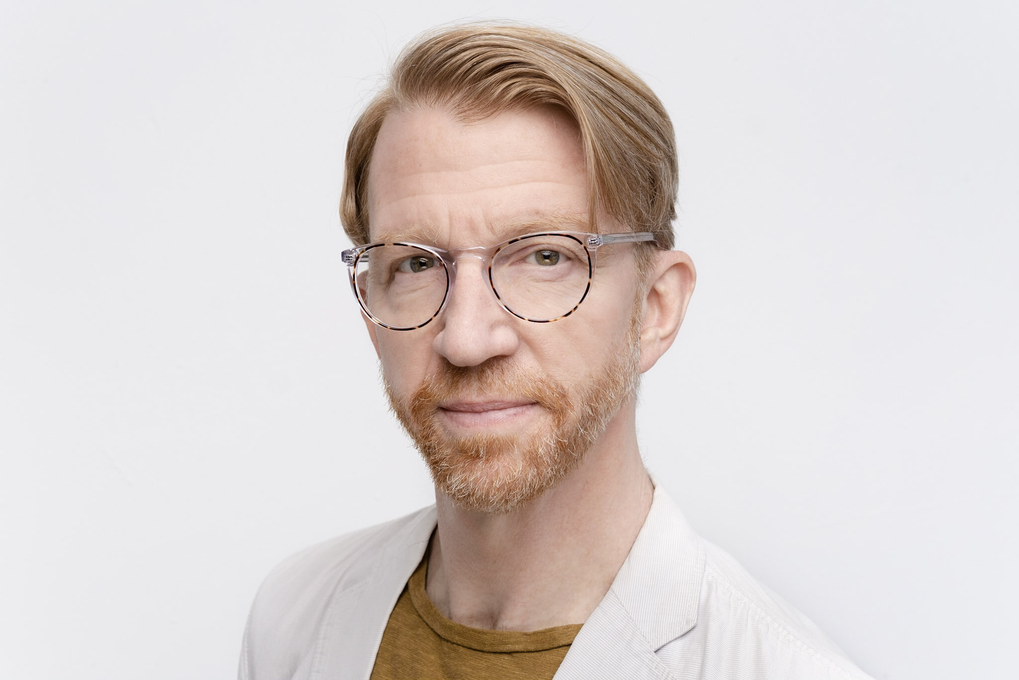 Photographer Francis Dzikovski, Portrait of a man with light skin, red hair, glasses, and a beard, wearing a beige blazer and brown shirt, standing against a white background.