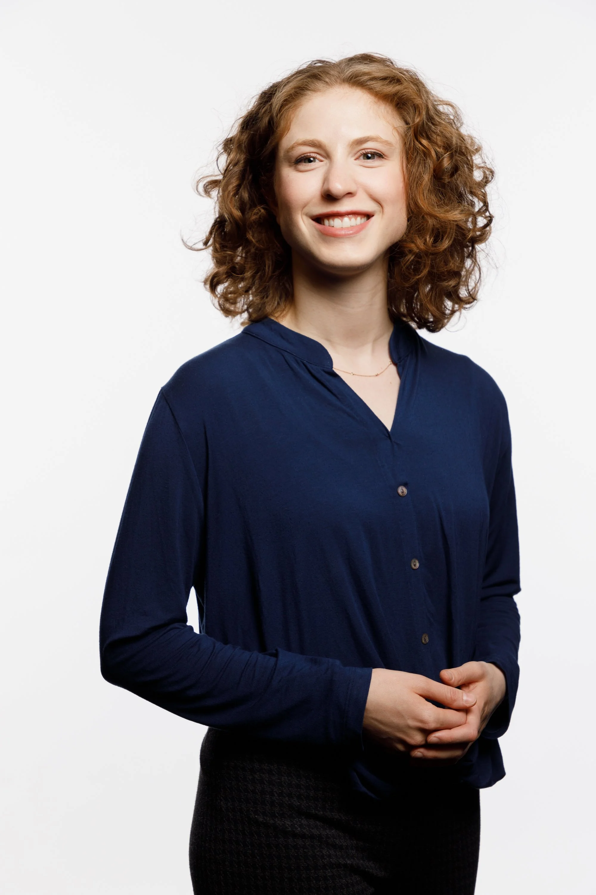 A woman with curly red hair wearing a navy blue blouse and black pants, standing against a plain white background and smiling.