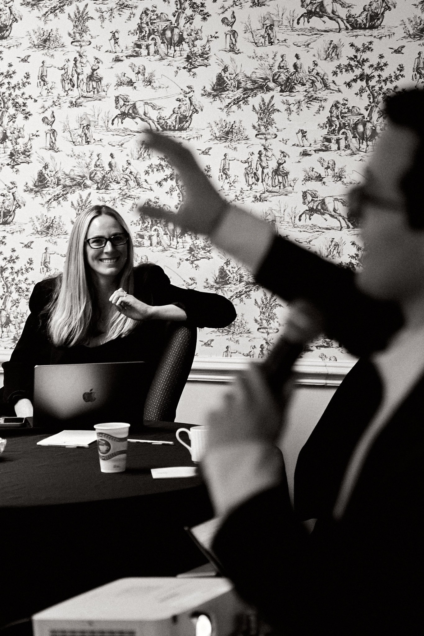 A woman with long blonde hair and glasses sitting at a meeting table, smiling, with a laptop in front of her. In the foreground, a person is gesturing with their hand while holding a mug. The background features a decorative wallpaper with a pastoral