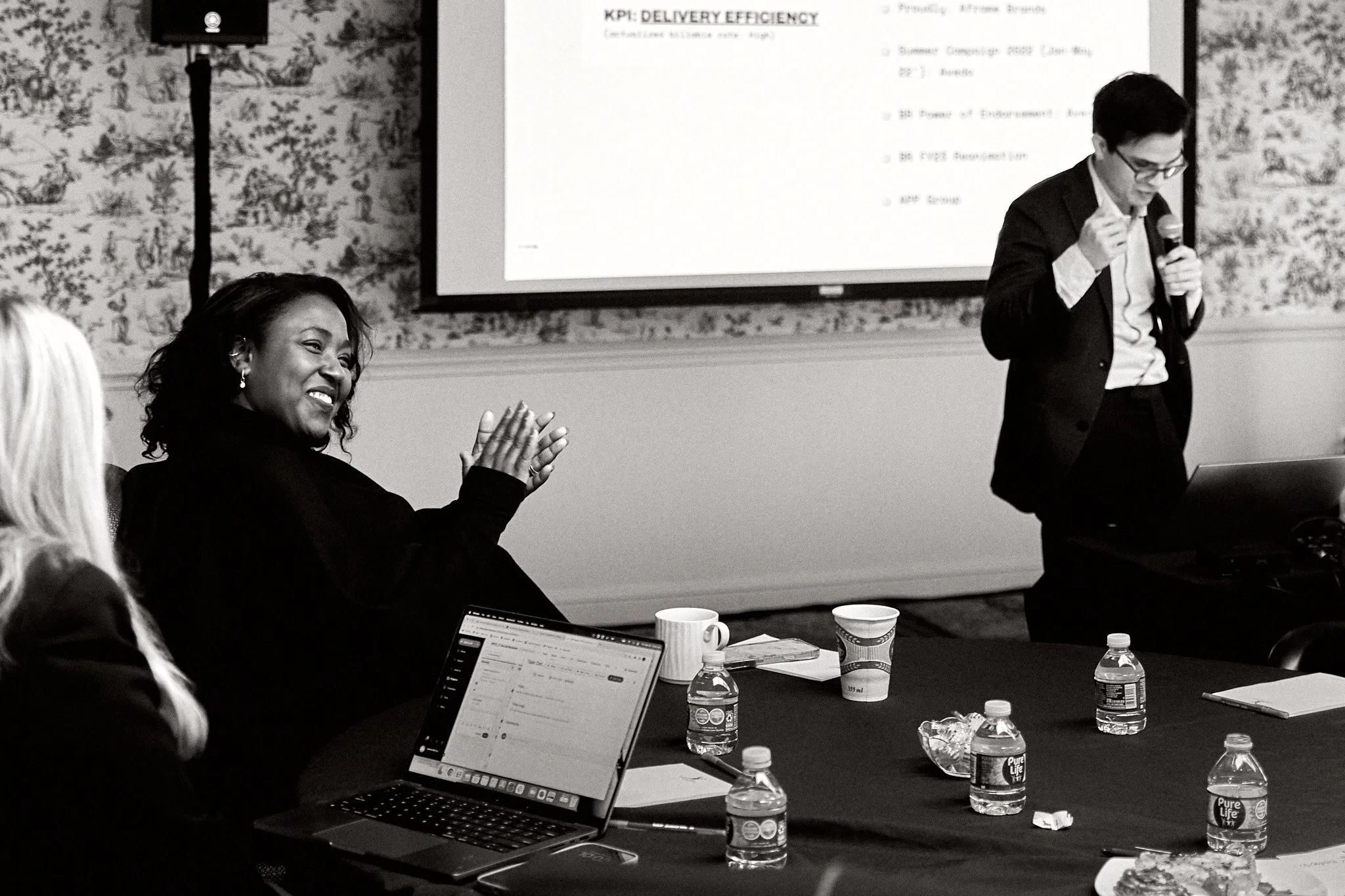 A woman is clapping and smiling during a presentation in a conference room. A man is standing, presenting with a microphone, and looking at his laptop. There are laptops, water bottles, and cups on the table, with a presentation slide projected on th