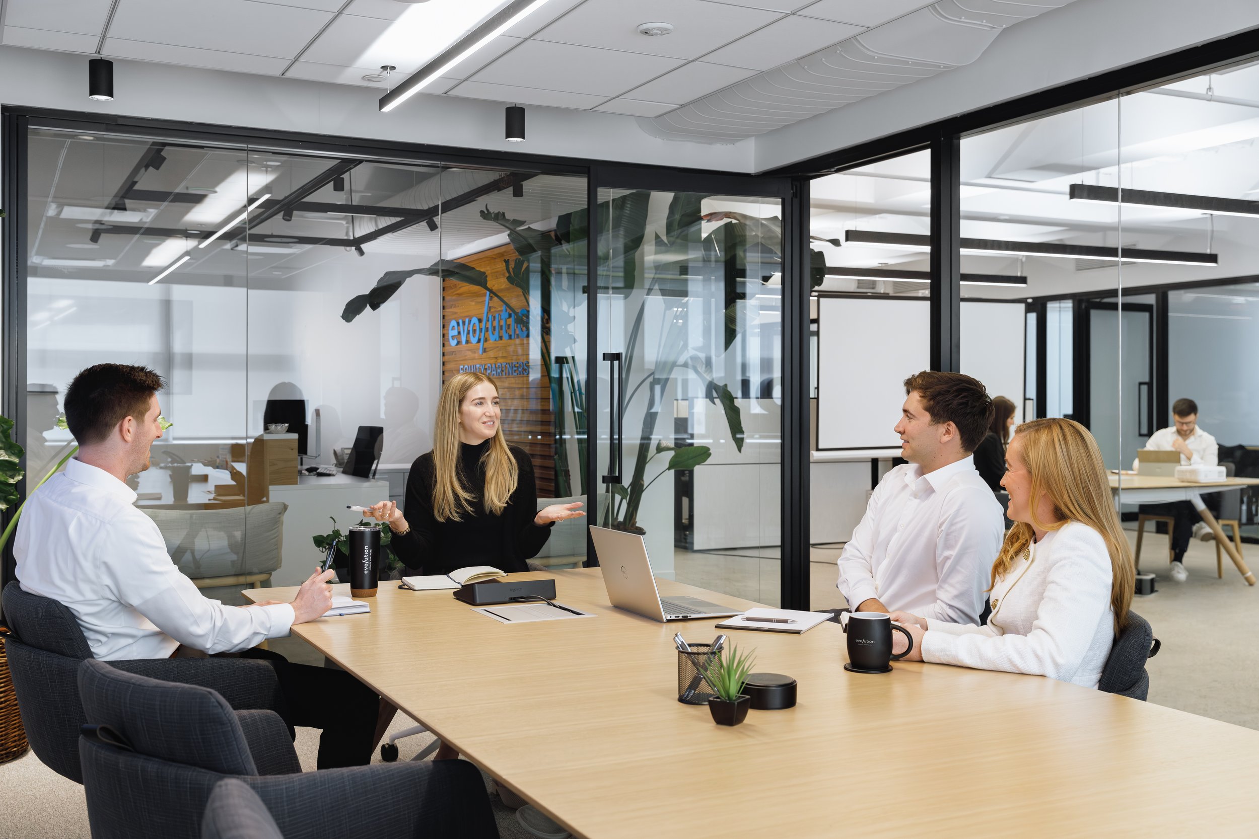 Four people in a business meeting in a modern conference room with glass walls, notebooks, laptops, and coffee mugs on the table.