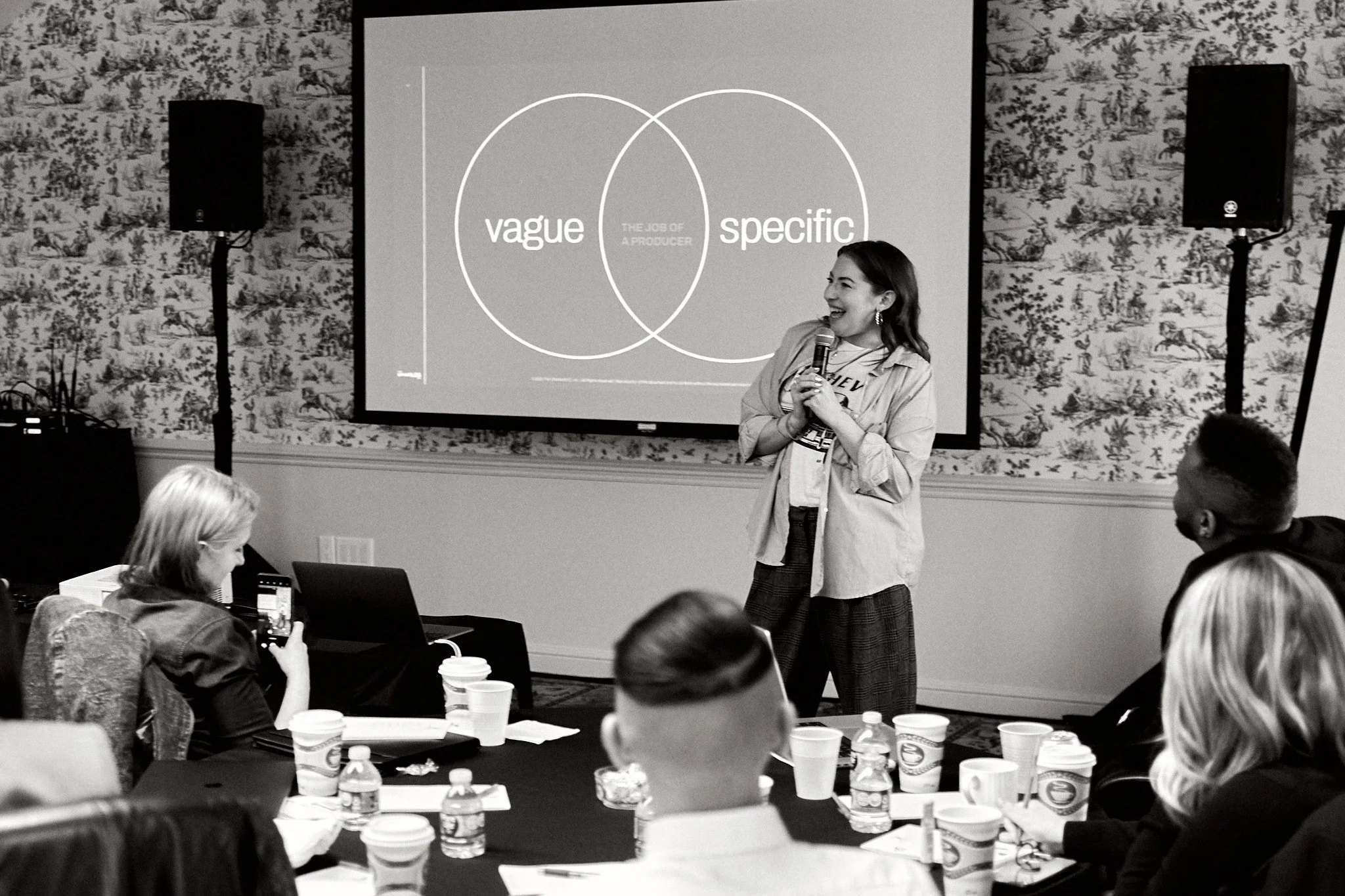 A woman giving a presentation in front of a projection screen showing a Venn diagram with the words "vague" and "specific" and the phrase "the job of a producer" in the overlapping area. Several audience members are seated at tables, some taking note