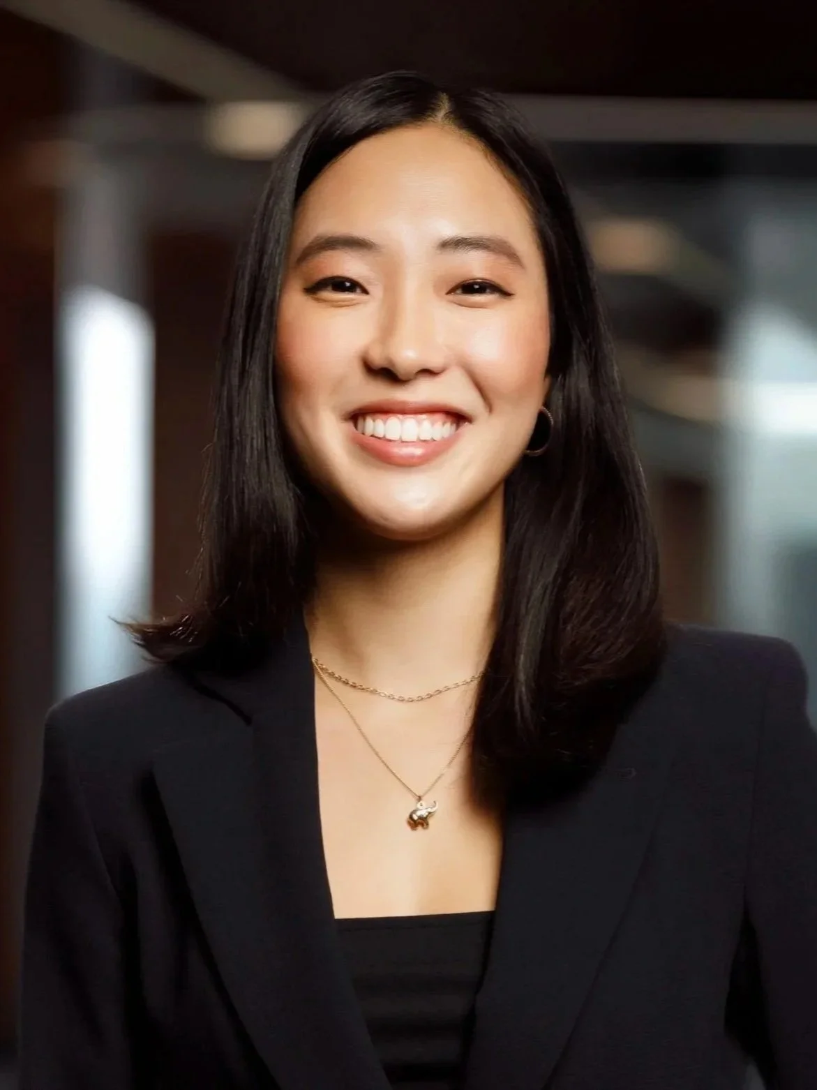 A smiling woman with black hair wearing a black blazer, gold necklace, and earrings, standing indoors with a blurred background.
