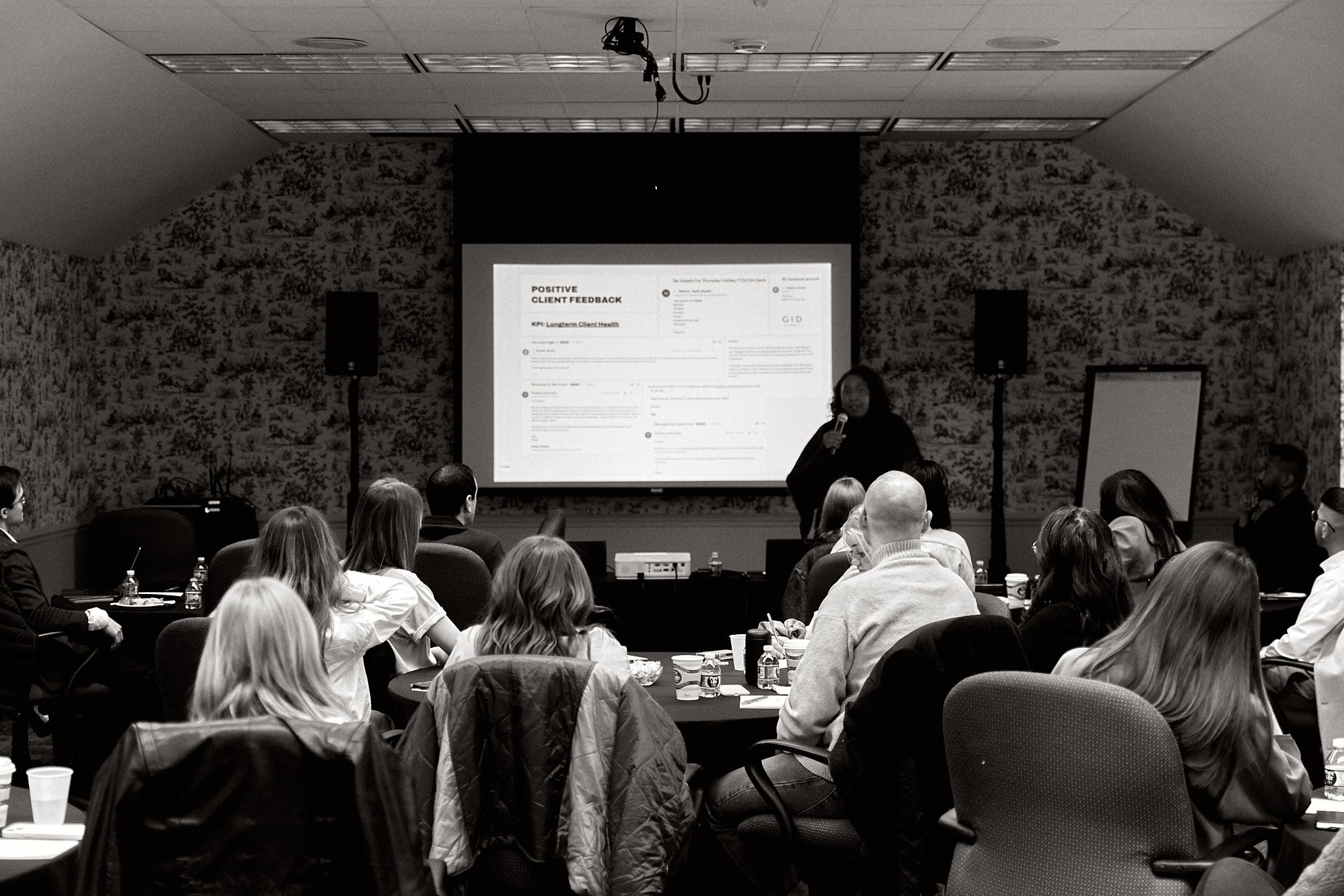 A woman giving a presentation to a group of people seated at round tables in a conference room with patterned wallpaper. The presenter stands near a large screen displaying slides, with a microphone in her hand.