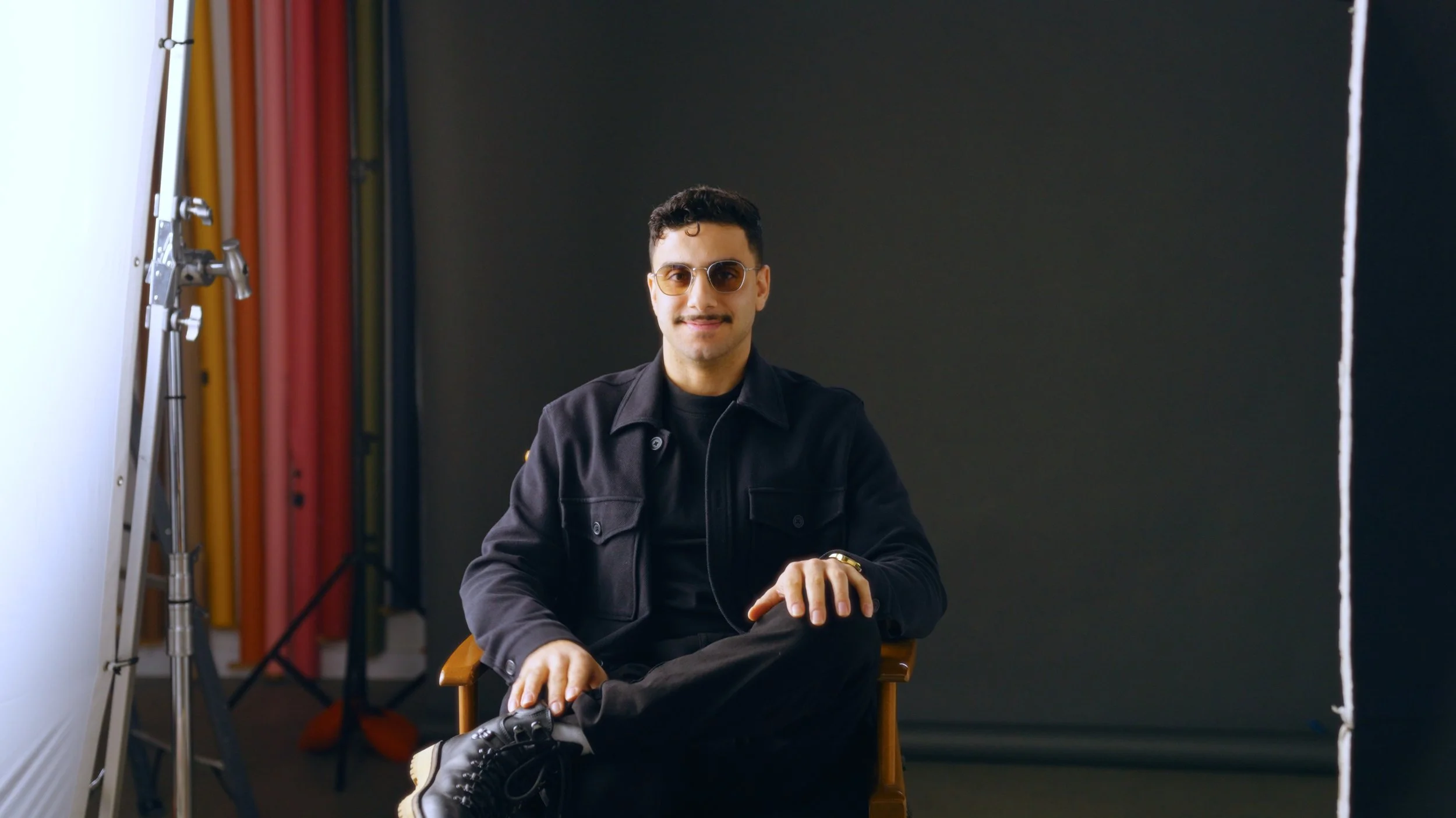 A young man sitting in a chair, smiling, wearing sunglasses, a black jacket, black pants, and black sneakers, with a dark backdrop and photography equipment in the background.
