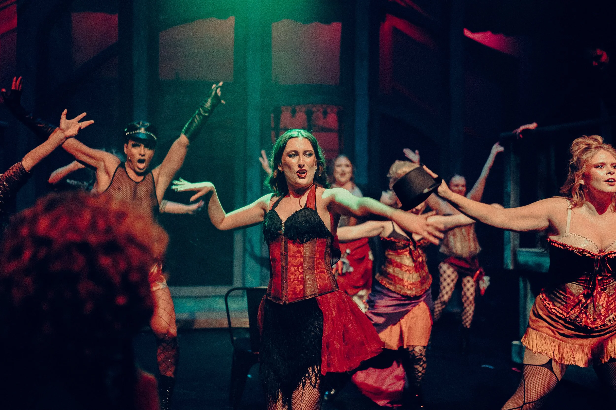 Performers in a theatrical dance, wearing corsets and fishnet stockings on stage under colorful lighting.