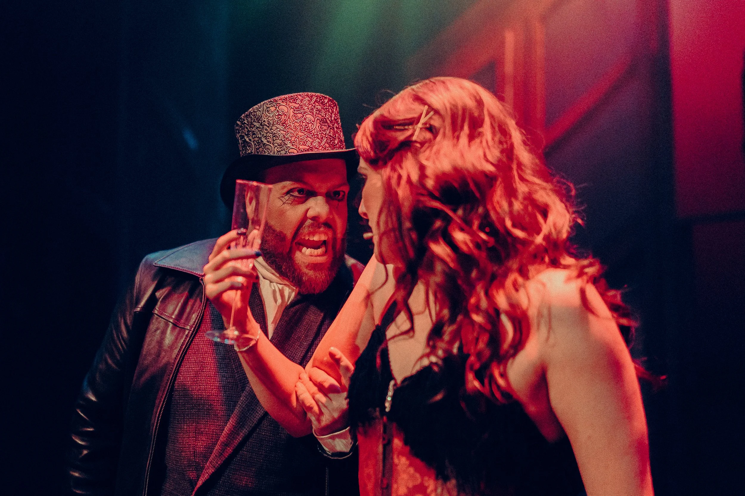 Two people in a dramatic, possibly theatrical, scene with intense expressions. One person wears a patterned top hat and holds a glass, appearing angry. The other person, with long curly hair, is engaged in the confrontation.