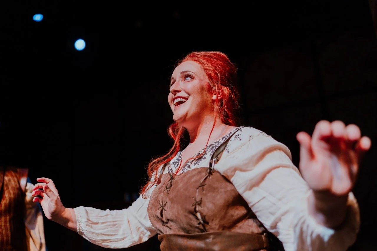 A person with red hair and a Renaissance-style costume smiling and gesturing on stage with a dark background.