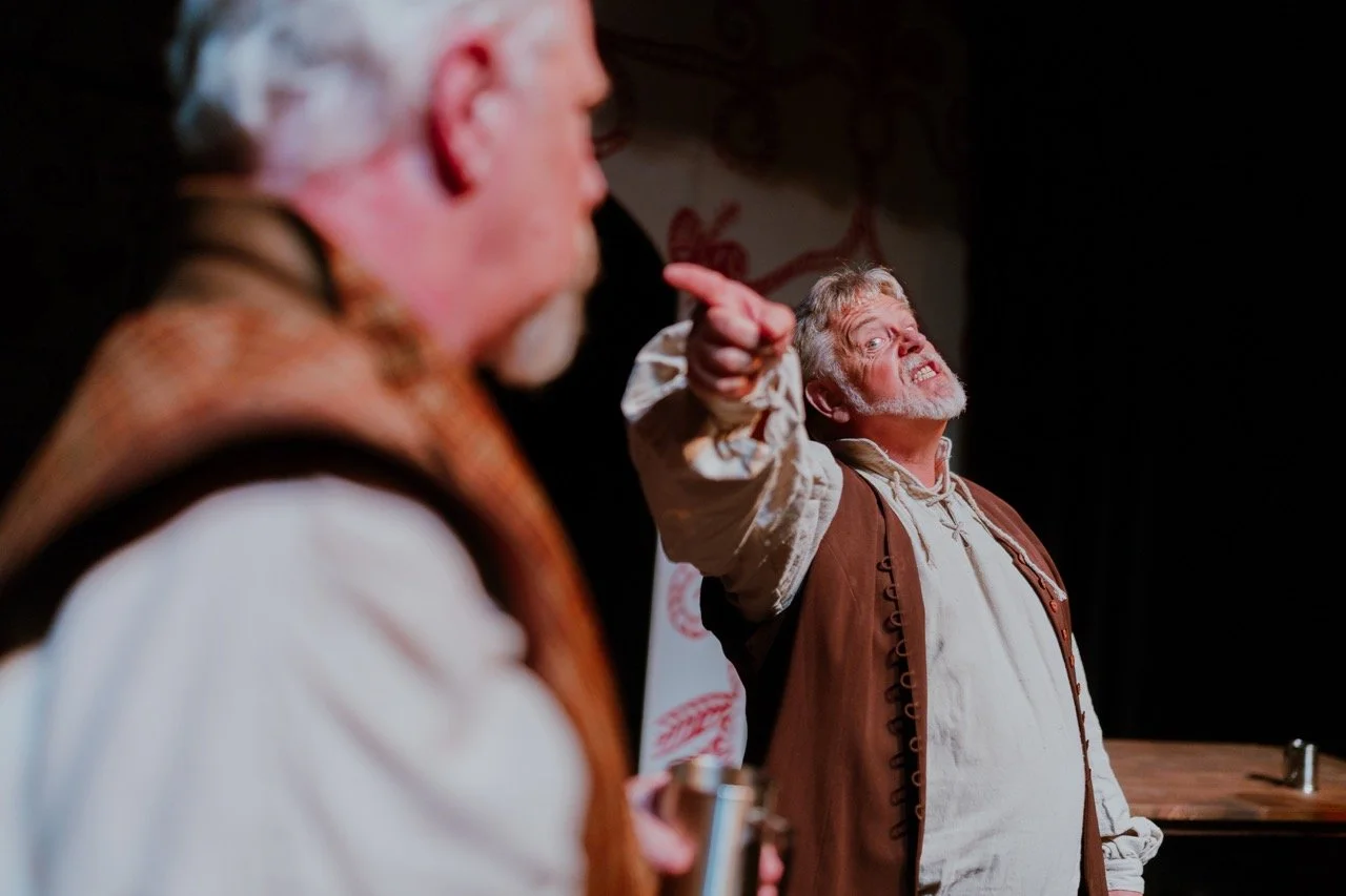 Two older men in period costumes engaged in a dramatic scene, with one man pointing passionately while the other holds a mug. The background is dimly lit, suggesting a stage setting.