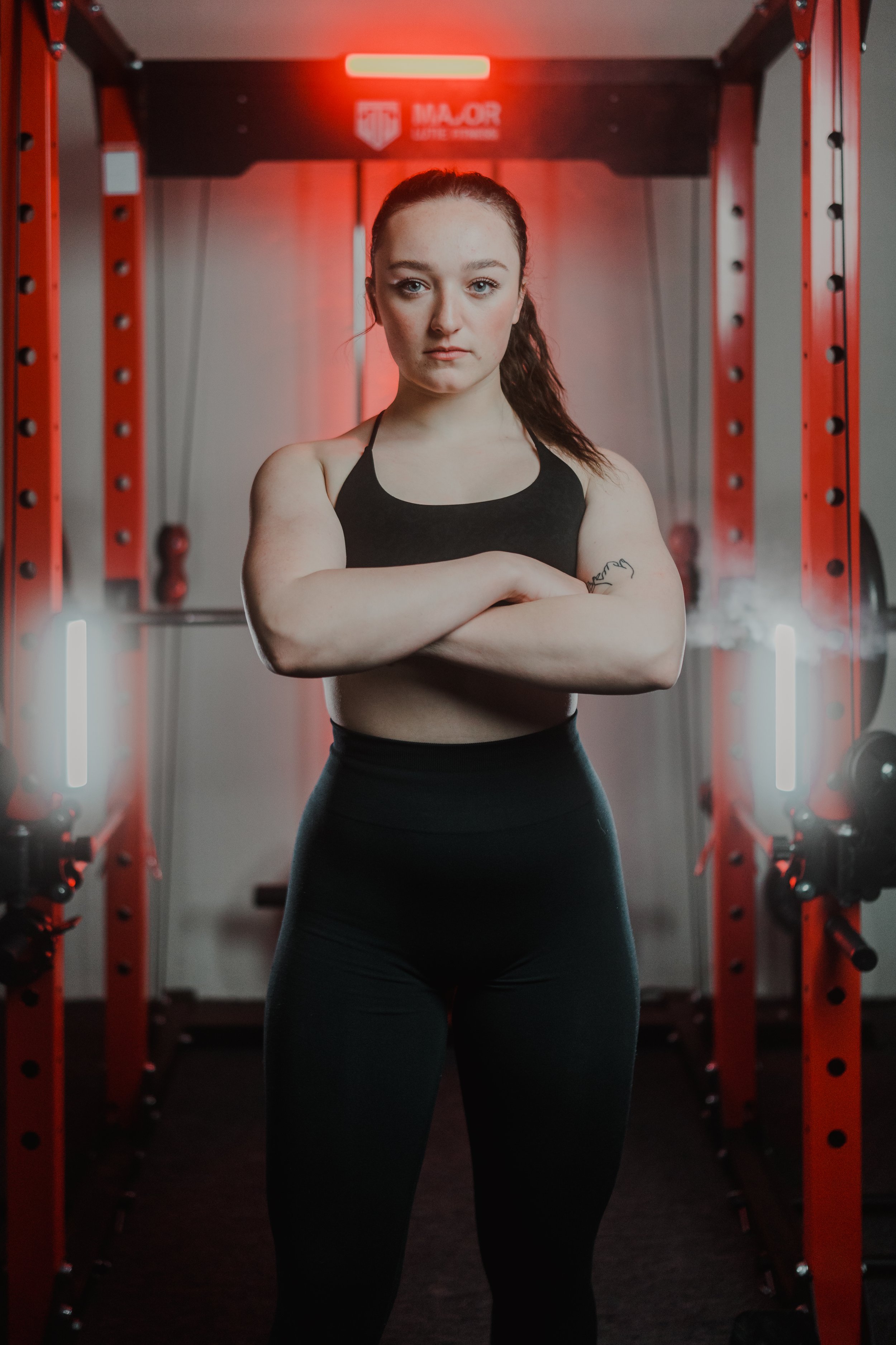 A female athlete standing confidently in a gym with red lighting and exercise equipment in the background.
