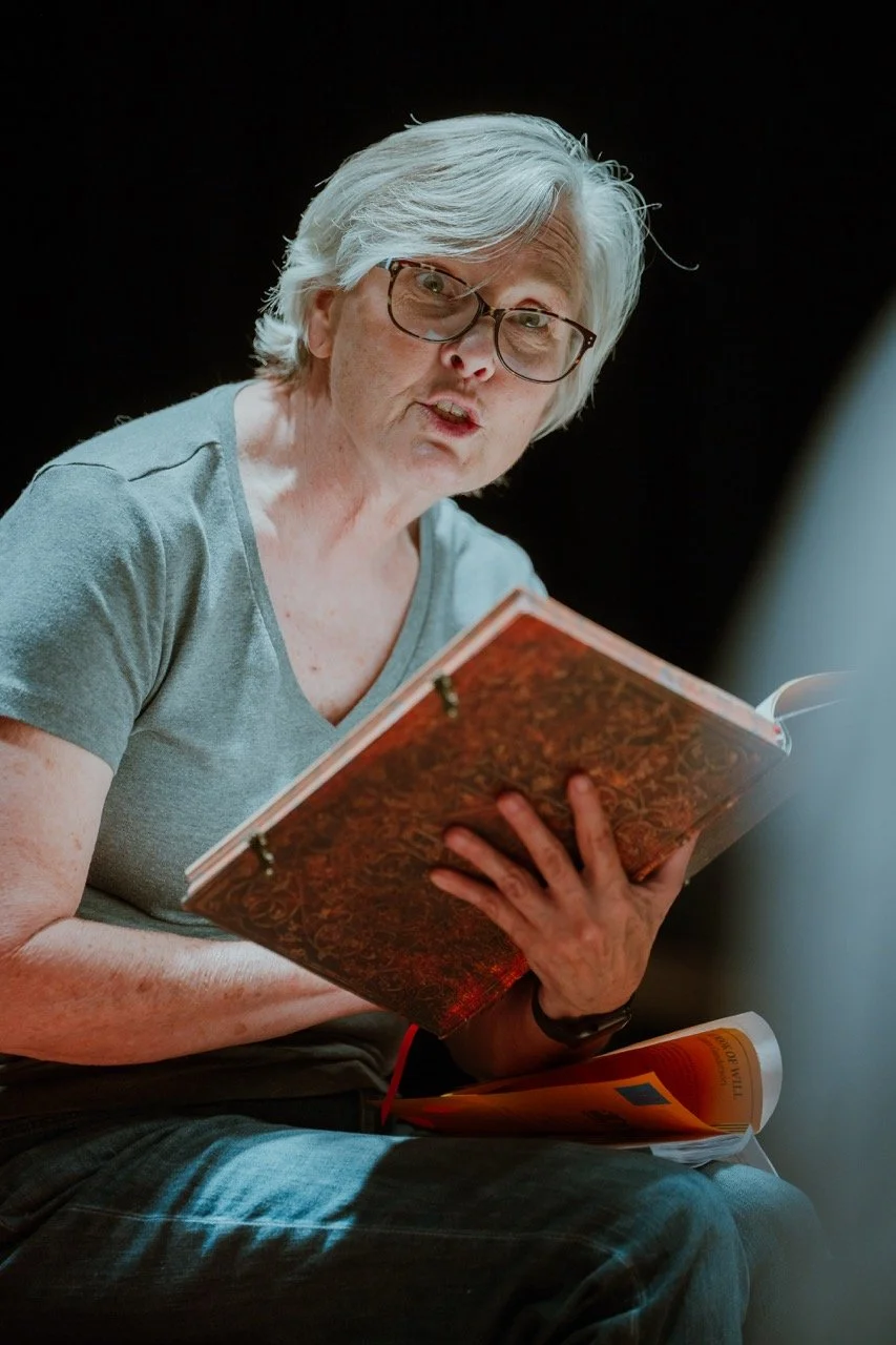 Older woman with glasses holding a book, reading or speaking passionately in a dimly lit setting.