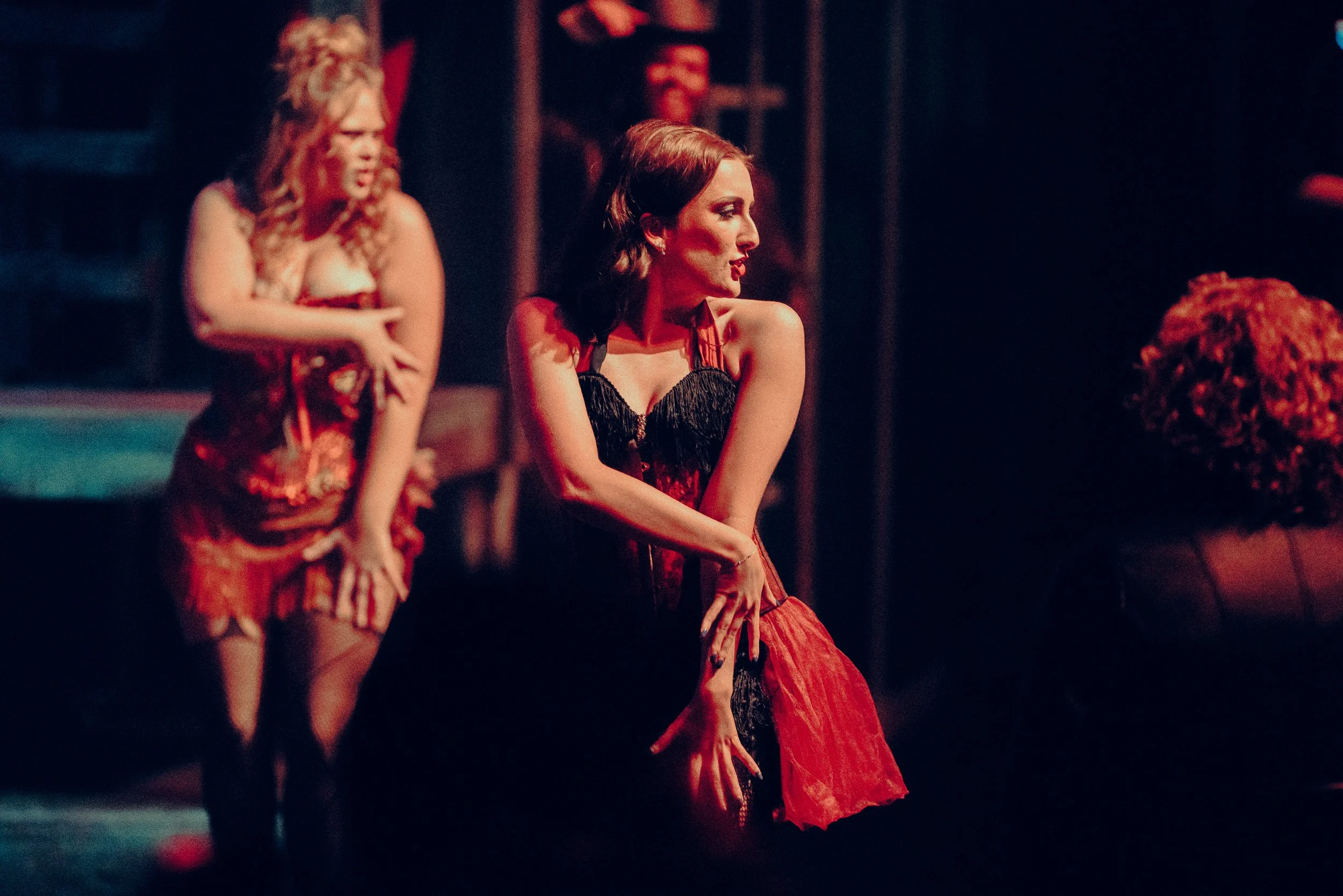 Two women dressed in burlesque-style costumes perform on stage with dramatic lighting.