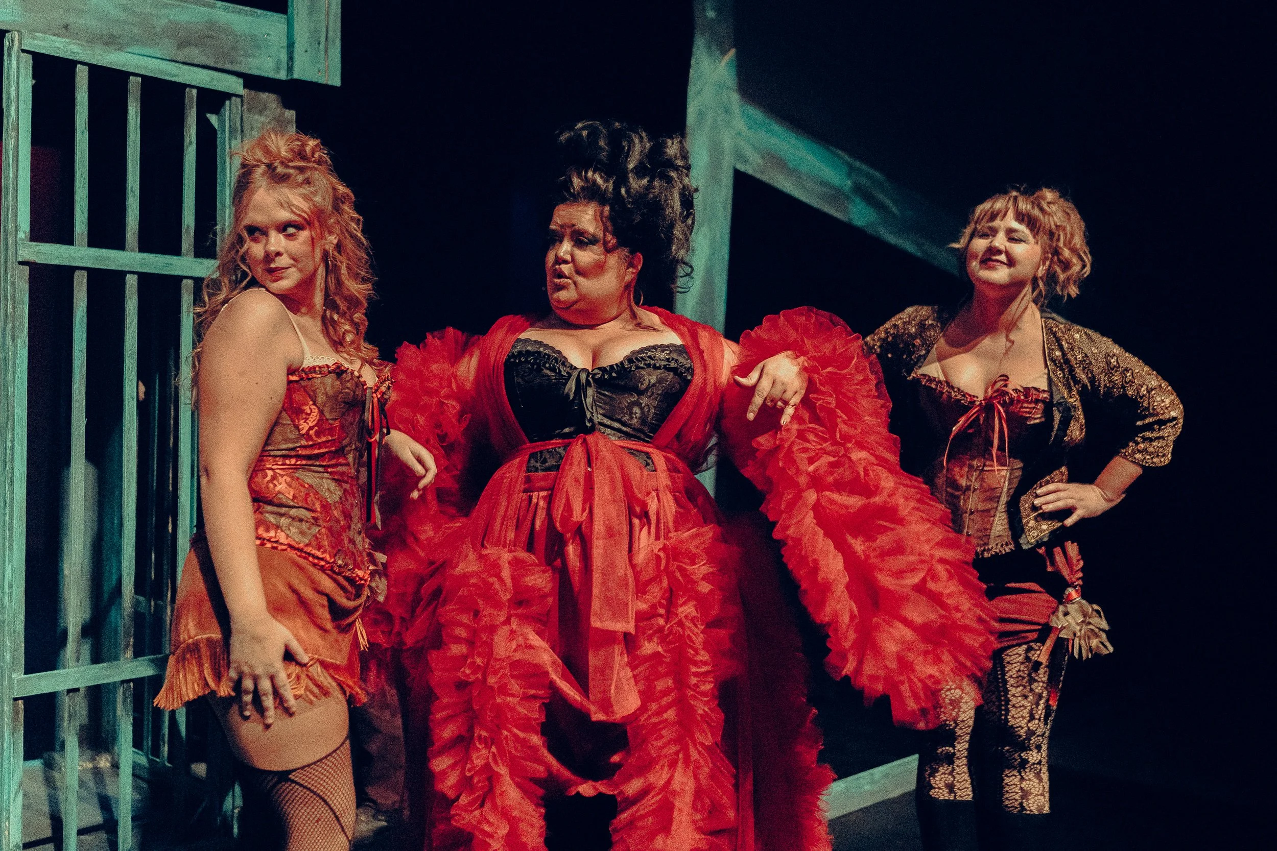 Three women in theatrical burlesque costumes stand on stage, posing confidently.