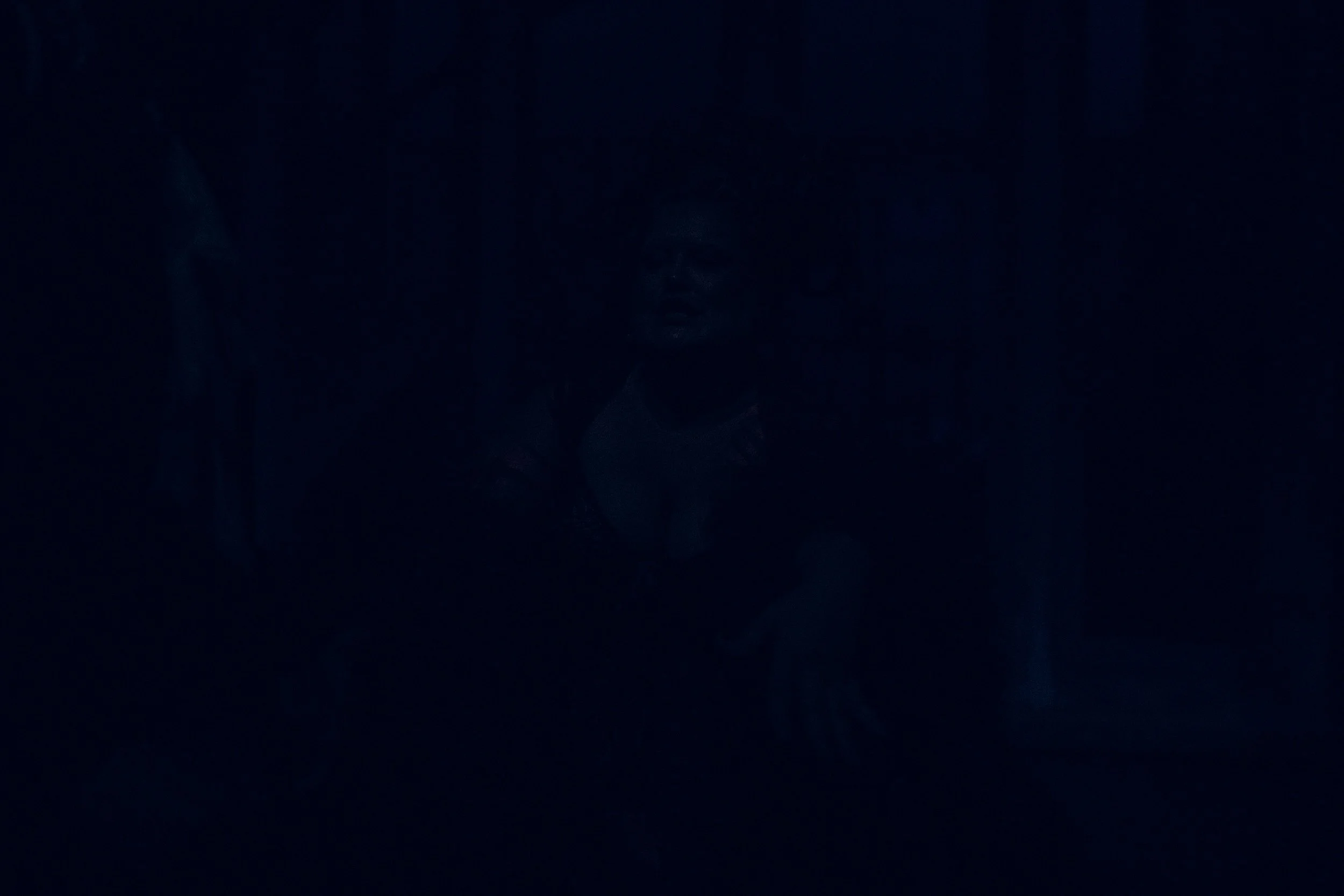A dimly lit scene with a person sitting down, barely visible due to low lighting.