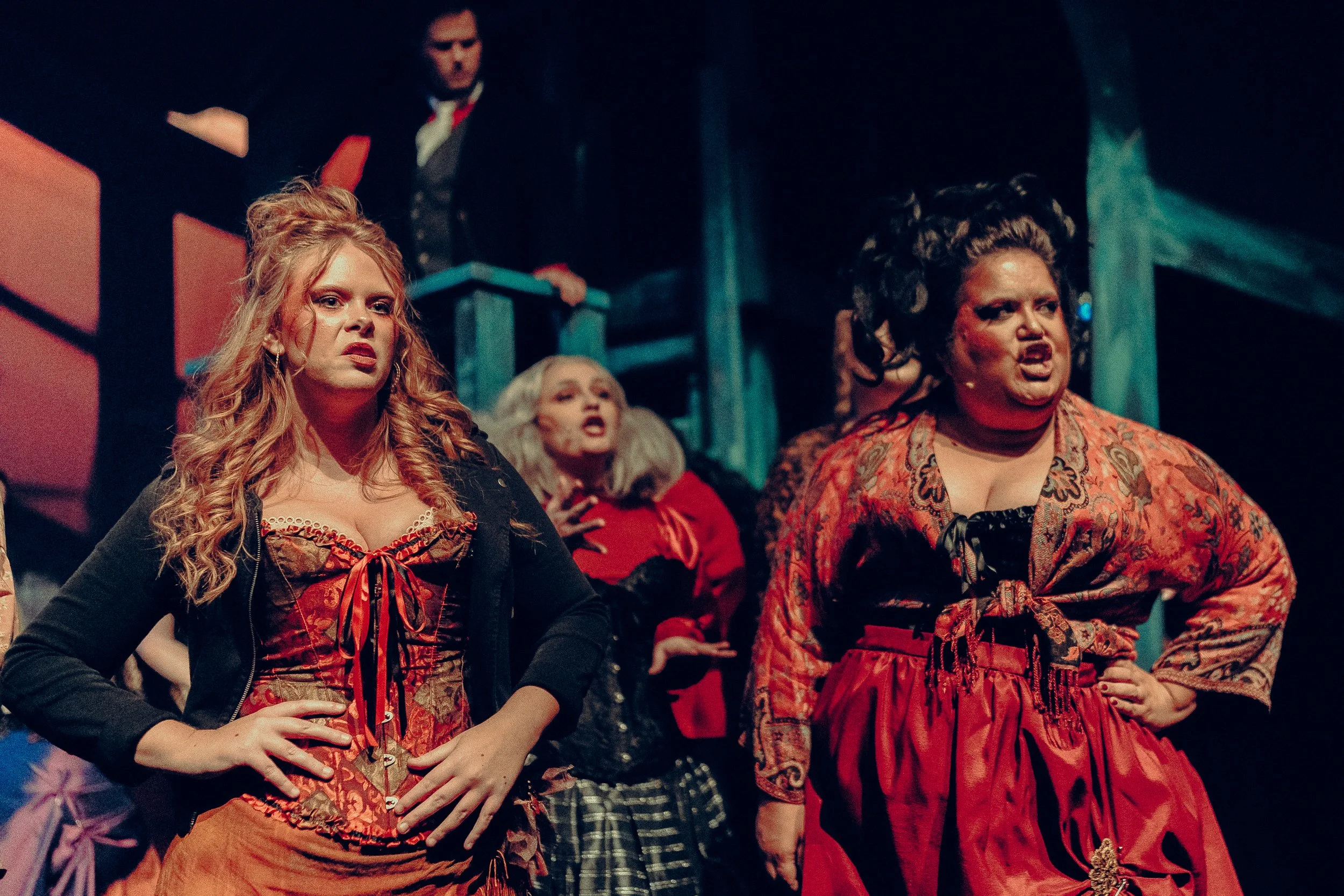 Stage performance with actors in period costumes, including two women in corsets and dresses, and a man in a suit, set in a theatrical setting.