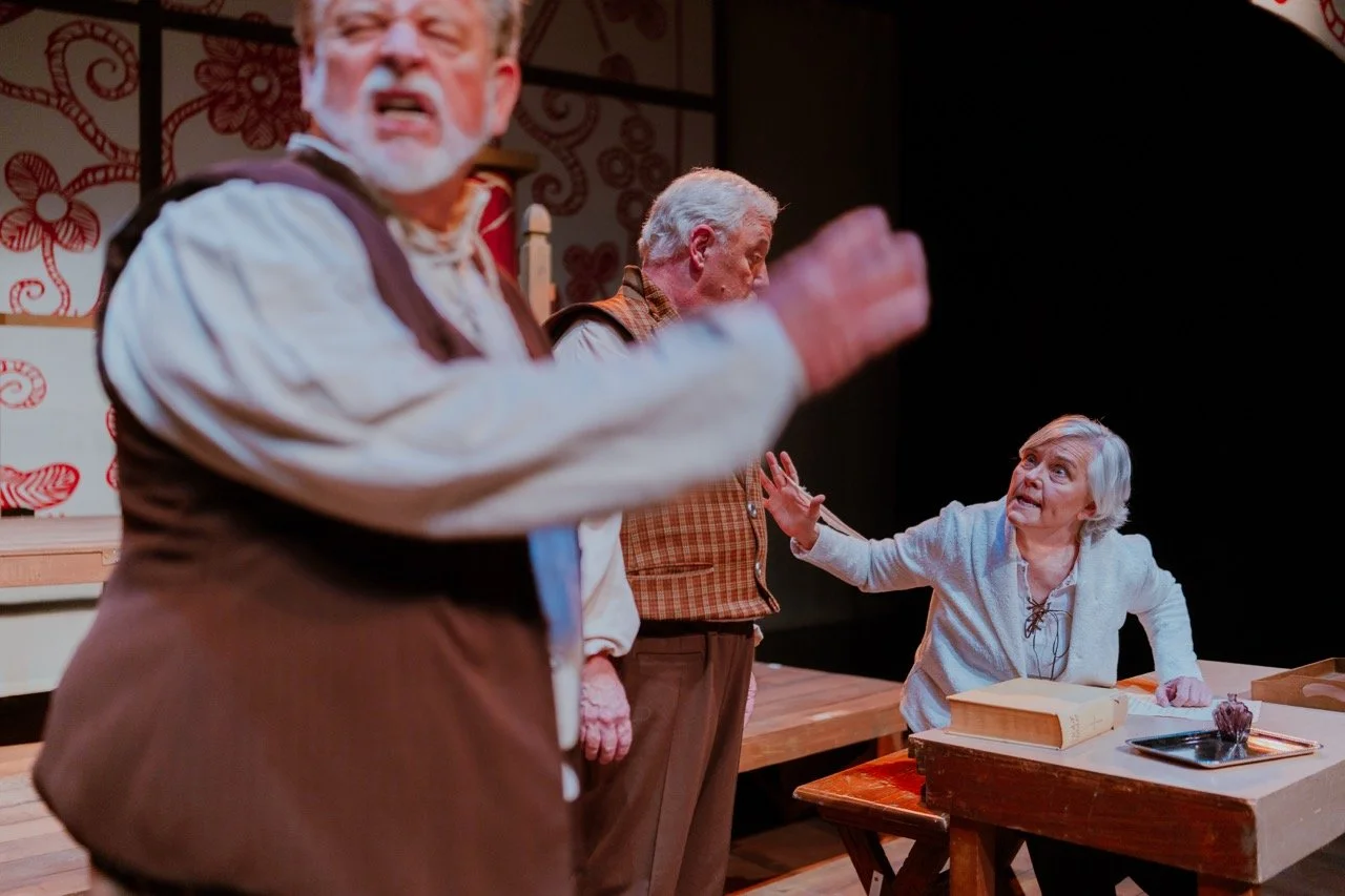 Three people performing in a theater play. Two elderly men are standing, one with an expressive face and arm raised, while an elderly woman sits at a table looking concerned. The setting appears to be historical or period-themed.