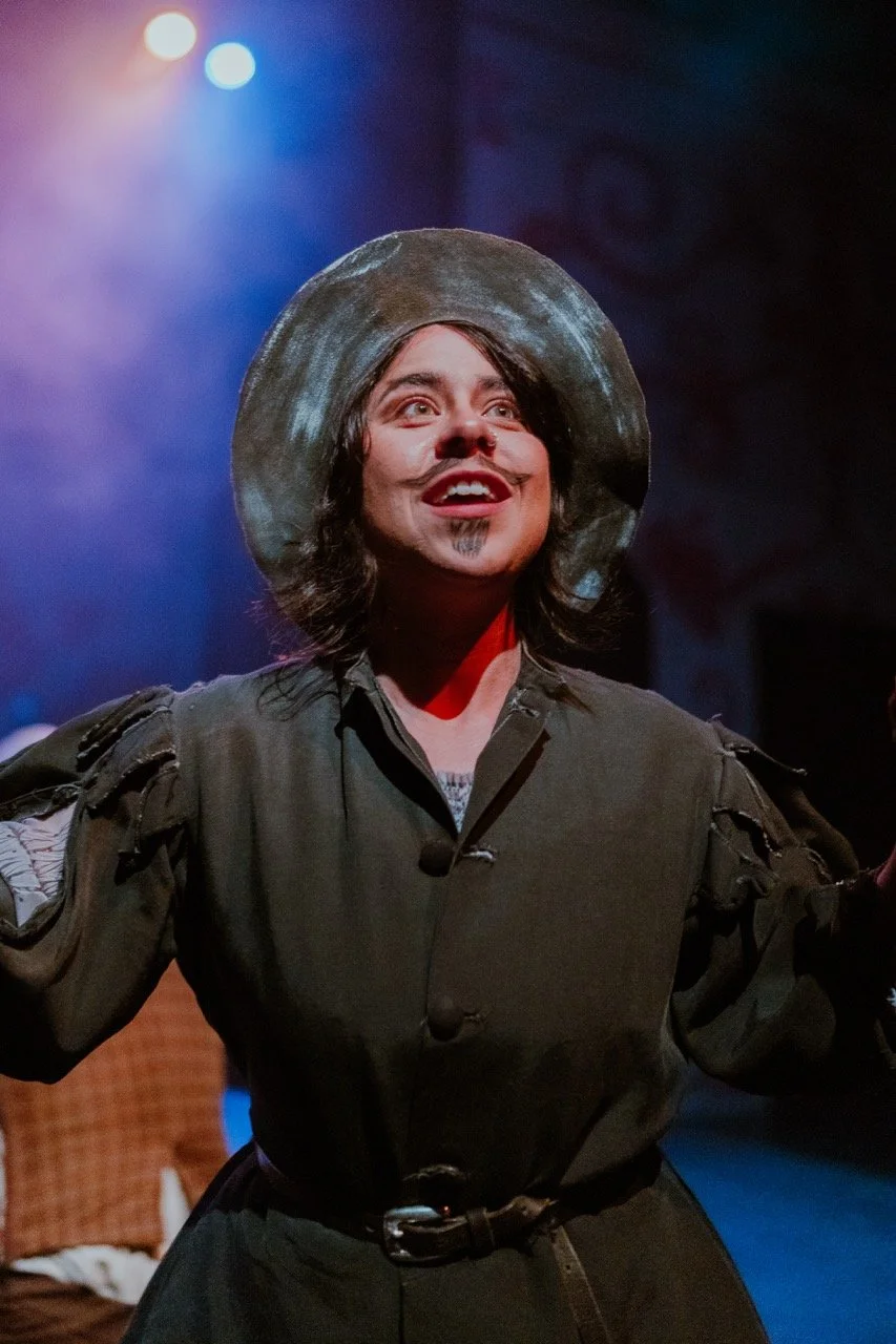 Person in theatrical costume with a hat and painted mustache, performing on stage with colorful lighting.