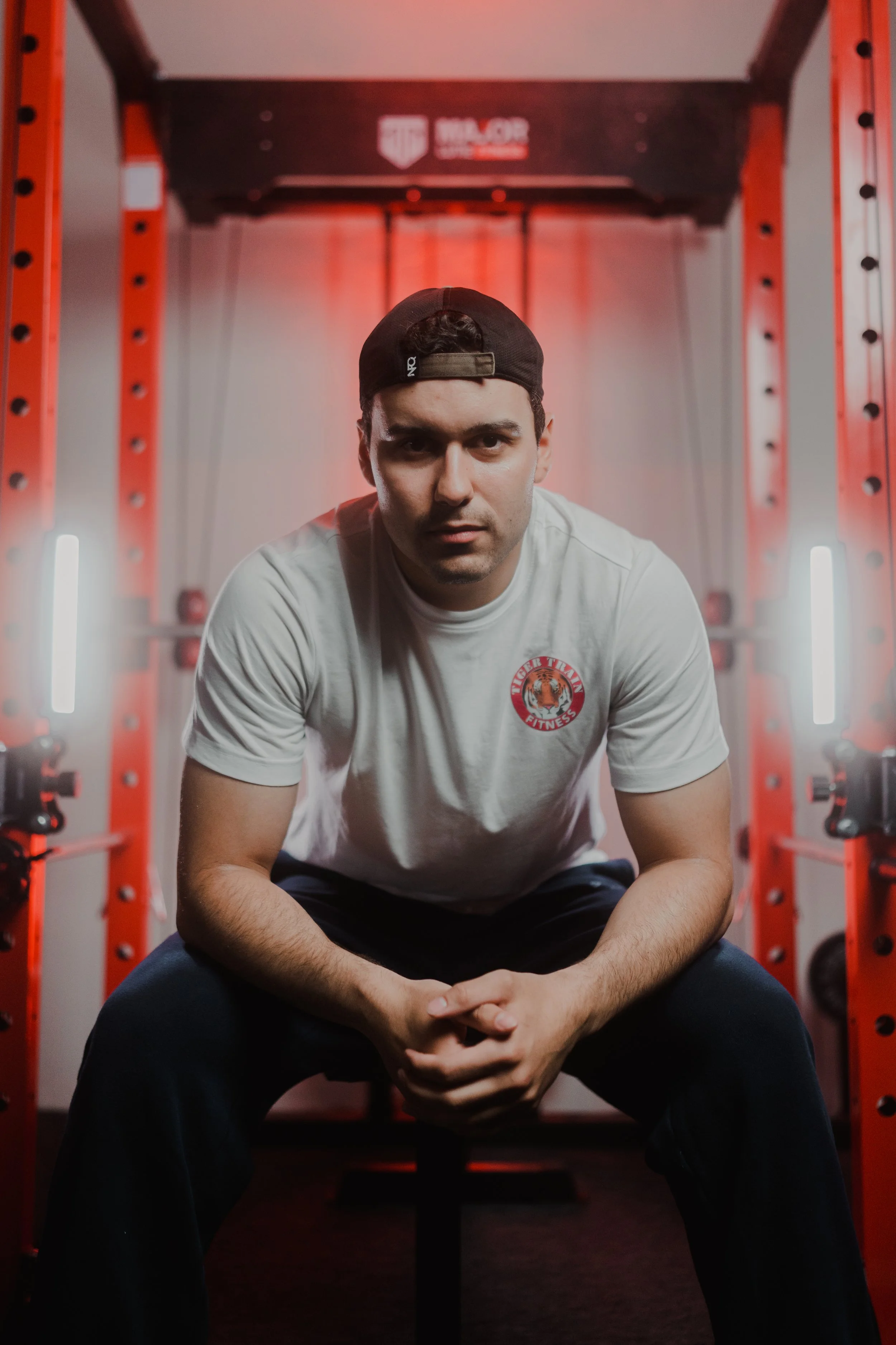 Man sitting in a gym with red equipment and wearing a white t-shirt and a black cap.