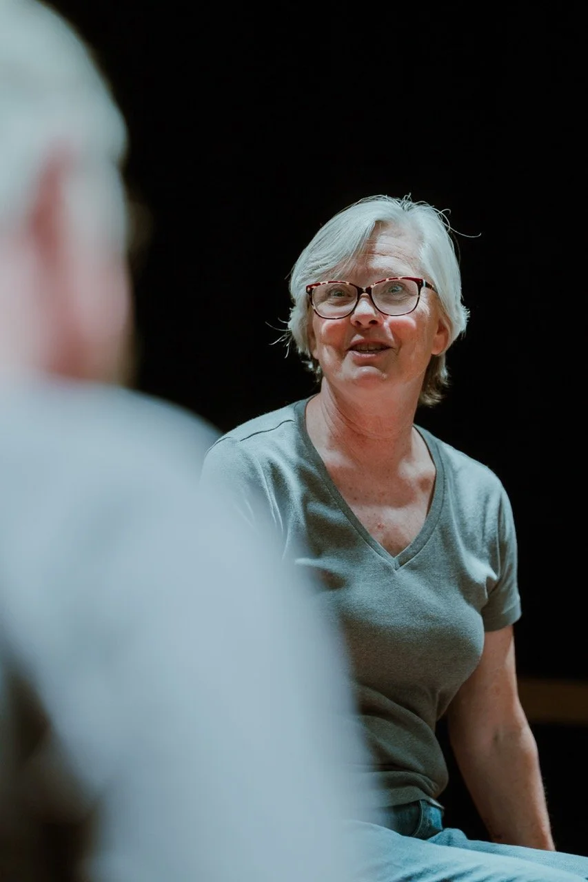 Older woman wearing glasses and a gray shirt, speaking or listening attentively in a dimly lit environment.