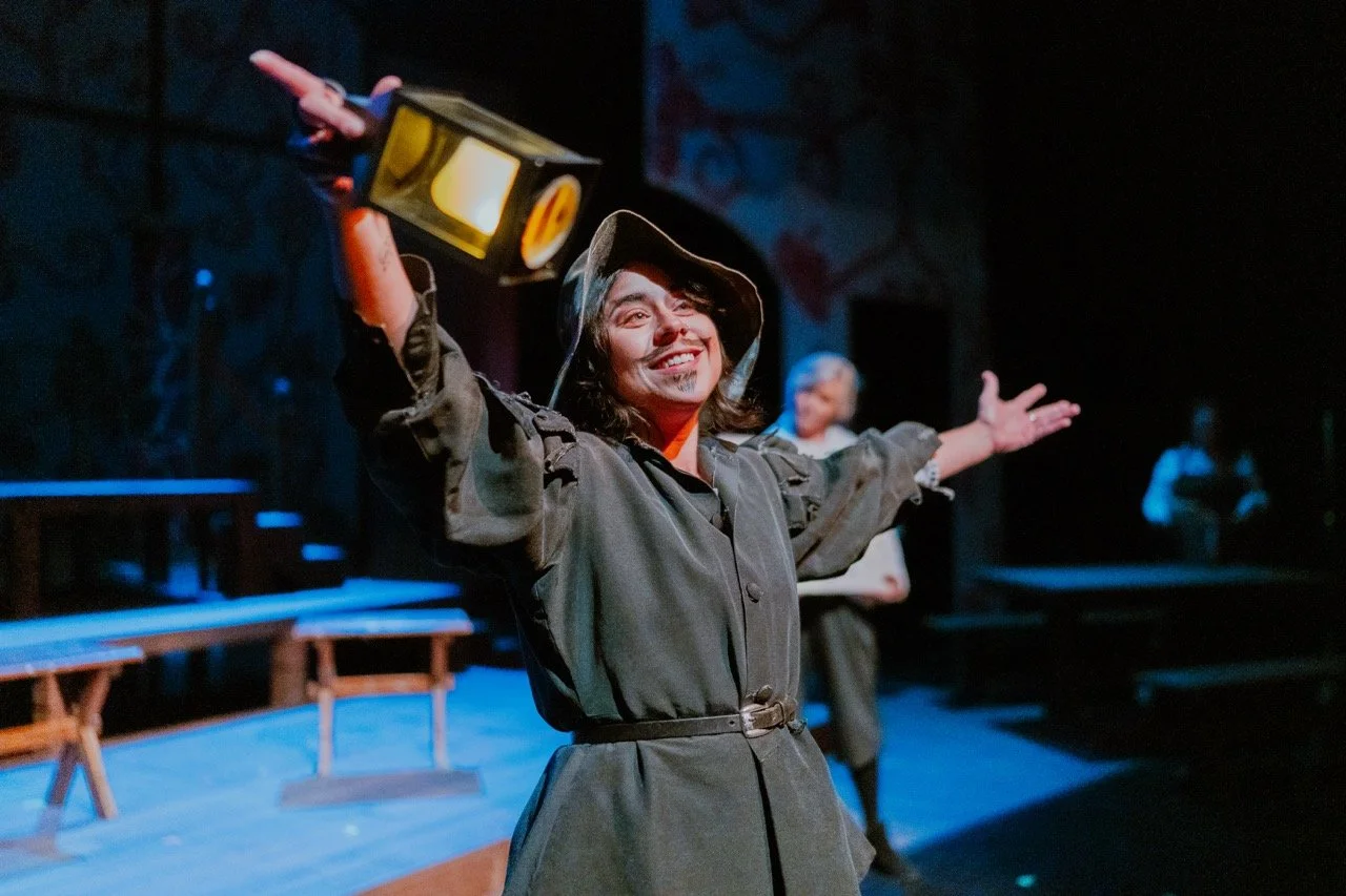 Person in costume on stage, holding a lantern, with arms outstretched, in a theatrical setting.