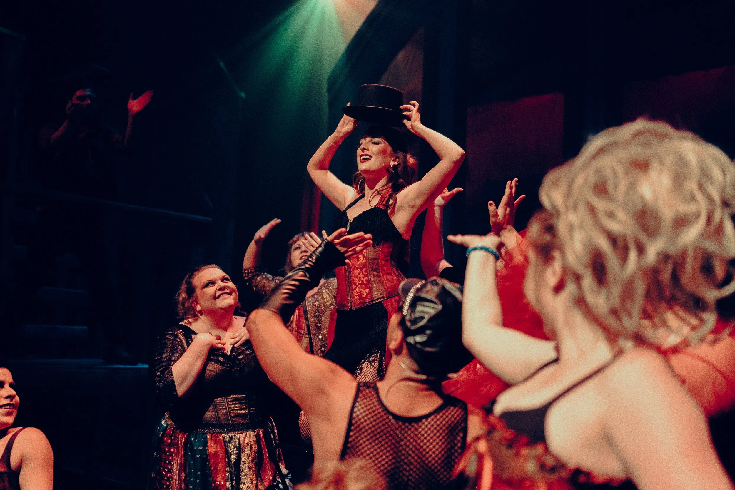 Performers in theatrical costumes on stage, one holding a top hat, with dramatic lighting.